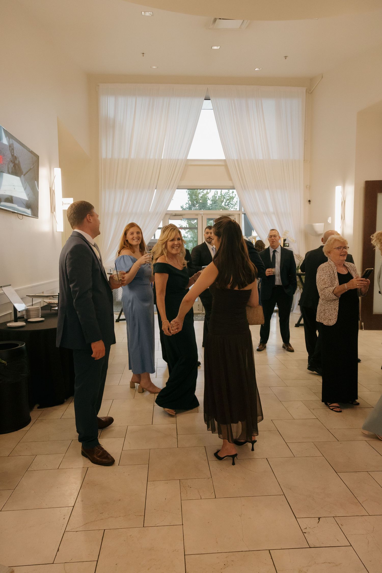 Wedding guests mingle in elegant venue lobby with white drapes and marble floors during reception.