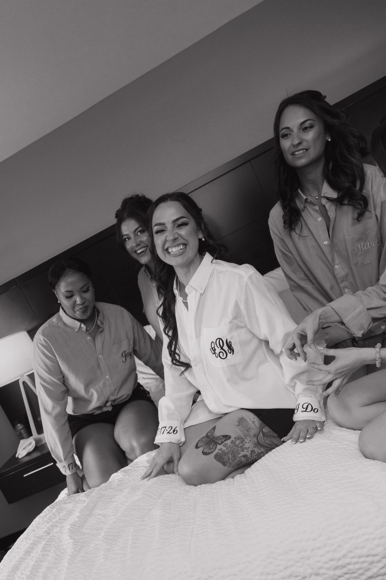 Bride and bridesmaids in monogrammed robes laughing together on a hotel bed during wedding morning preparations.