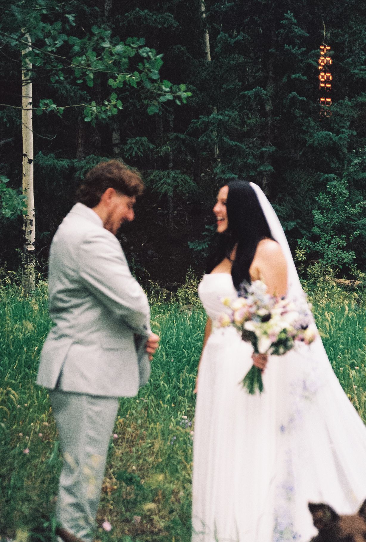 Bride in white gown holding a floral bouquet laughs joyfully with groom during their first look in a lush forest setting.