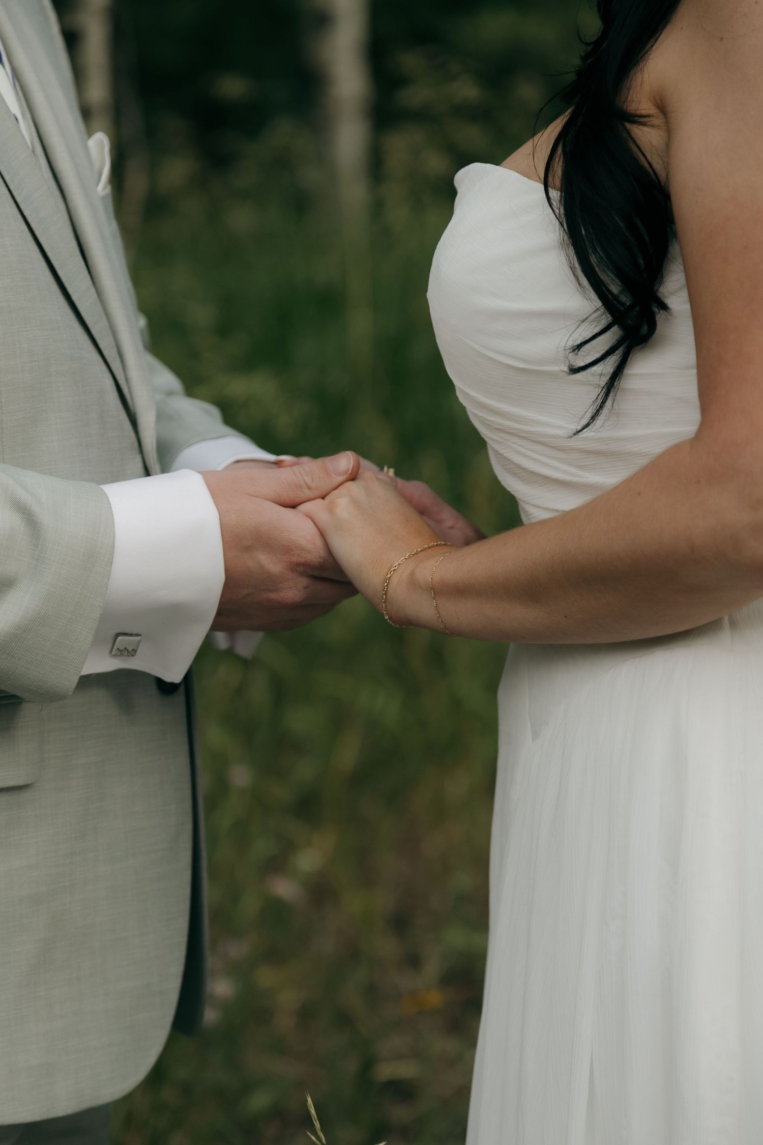Close-up of bride and groom holding hands during outdoor wedding ceremony, bride wearing gold bracelet.