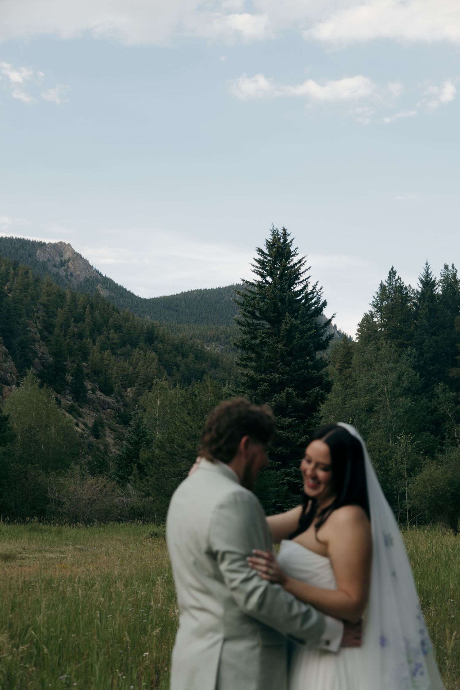 Bride and groom embracing in a meadow with mountain and pine tree backdrop on wedding day.