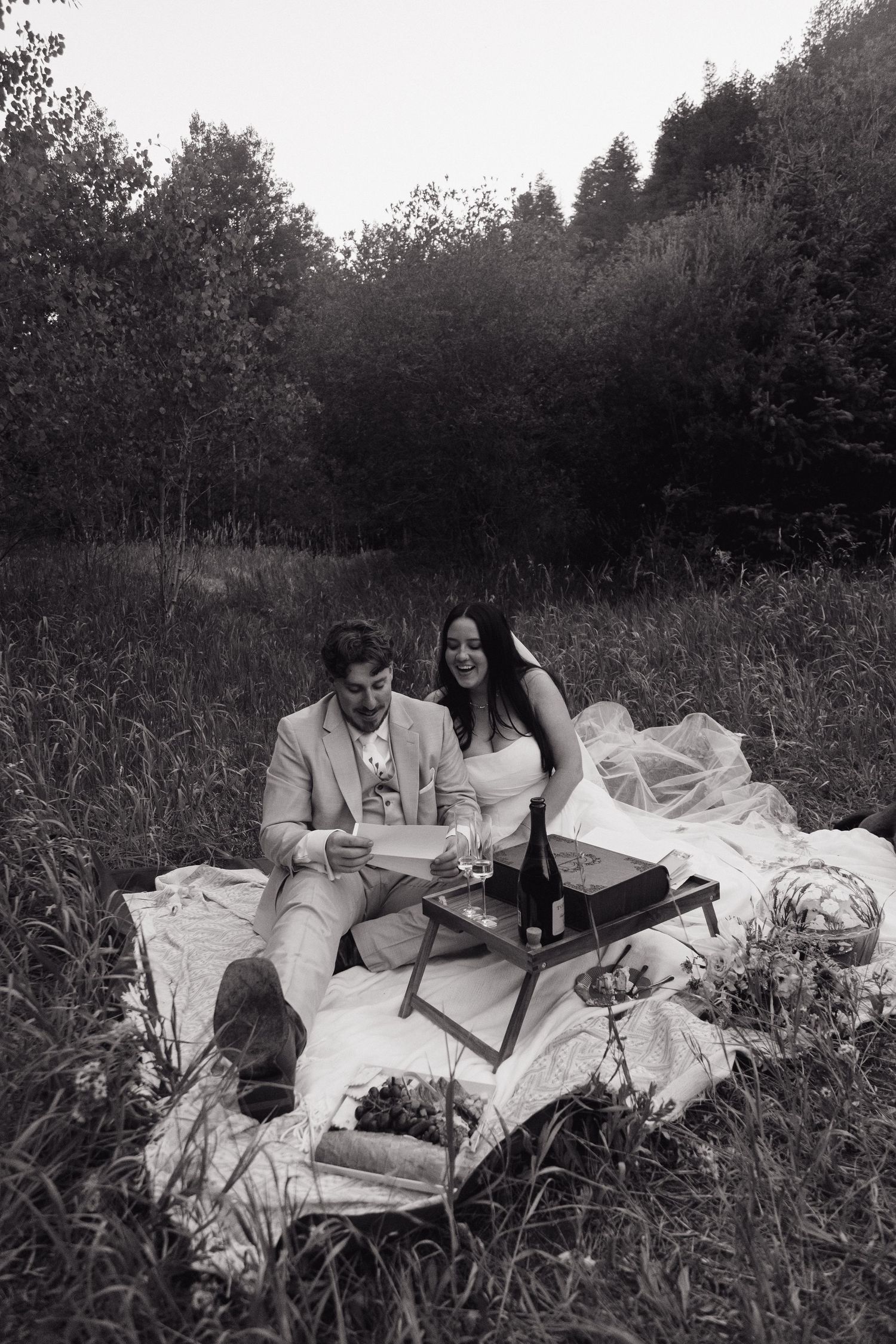 Black and white photo of bride and groom relaxing at outdoor picnic in forest meadow.