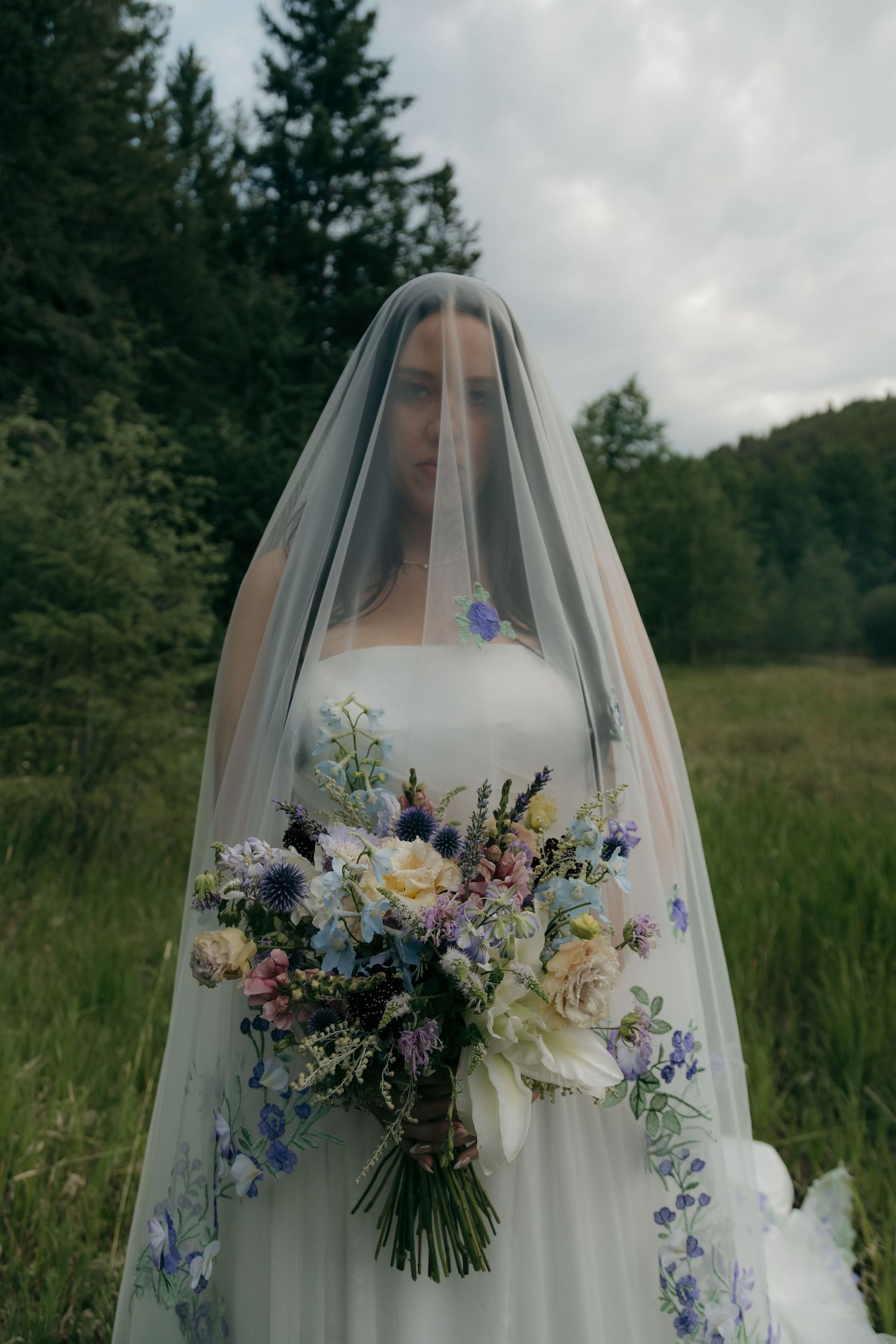 Elegant bride wearing floral embroidered veil holds a colorful wildflower bouquet in a forest meadow setting.