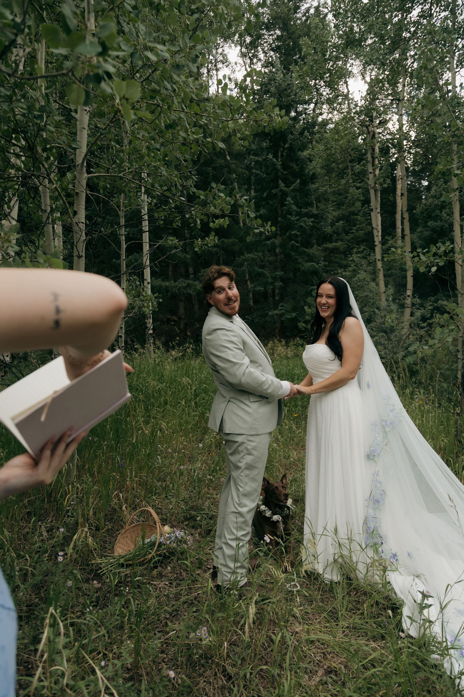 Smiling bride and groom hold hands during intimate forest elopement ceremony with officiant reading vows nearby.