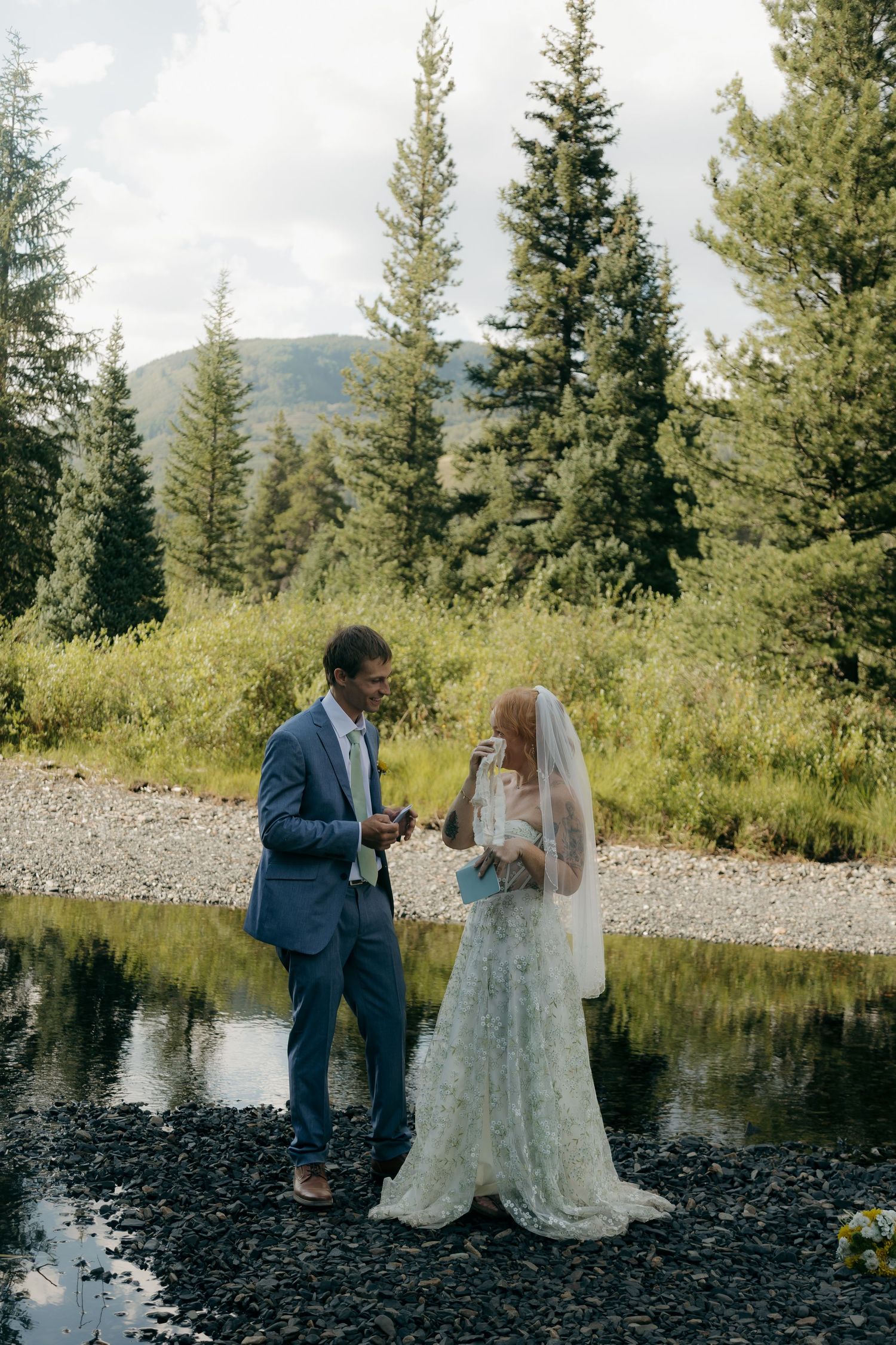 Bride and groom toasting with champagne beside a mountain stream surrounded by evergreen trees.