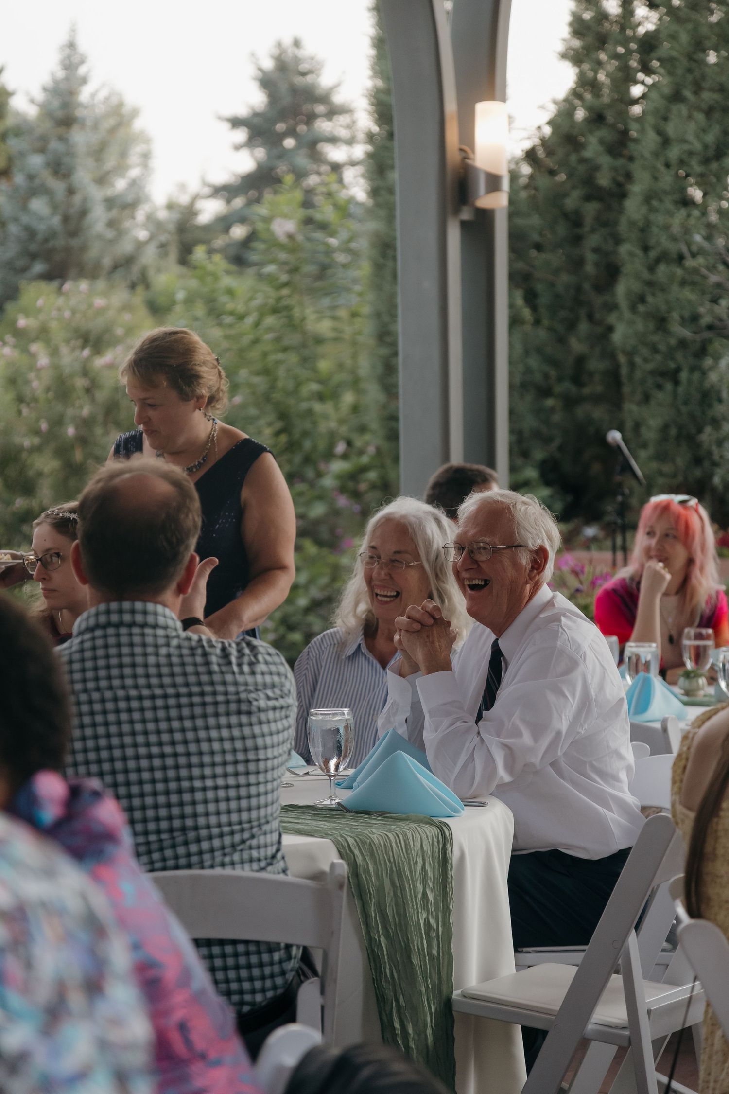 Elderly wedding guests laugh joyfully at outdoor reception dinner table with green decor.