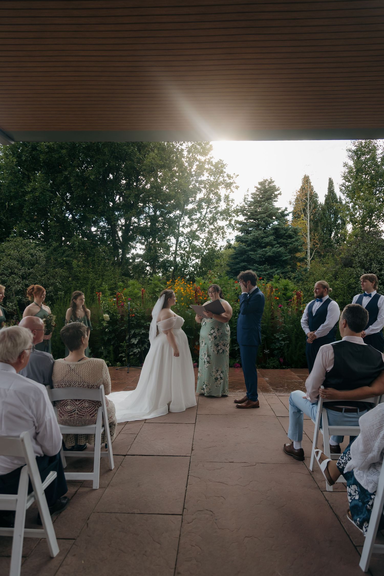 Bride and groom exchange vows under a pavilion at sunset, surrounded by wedding party and seated guests in a garden.