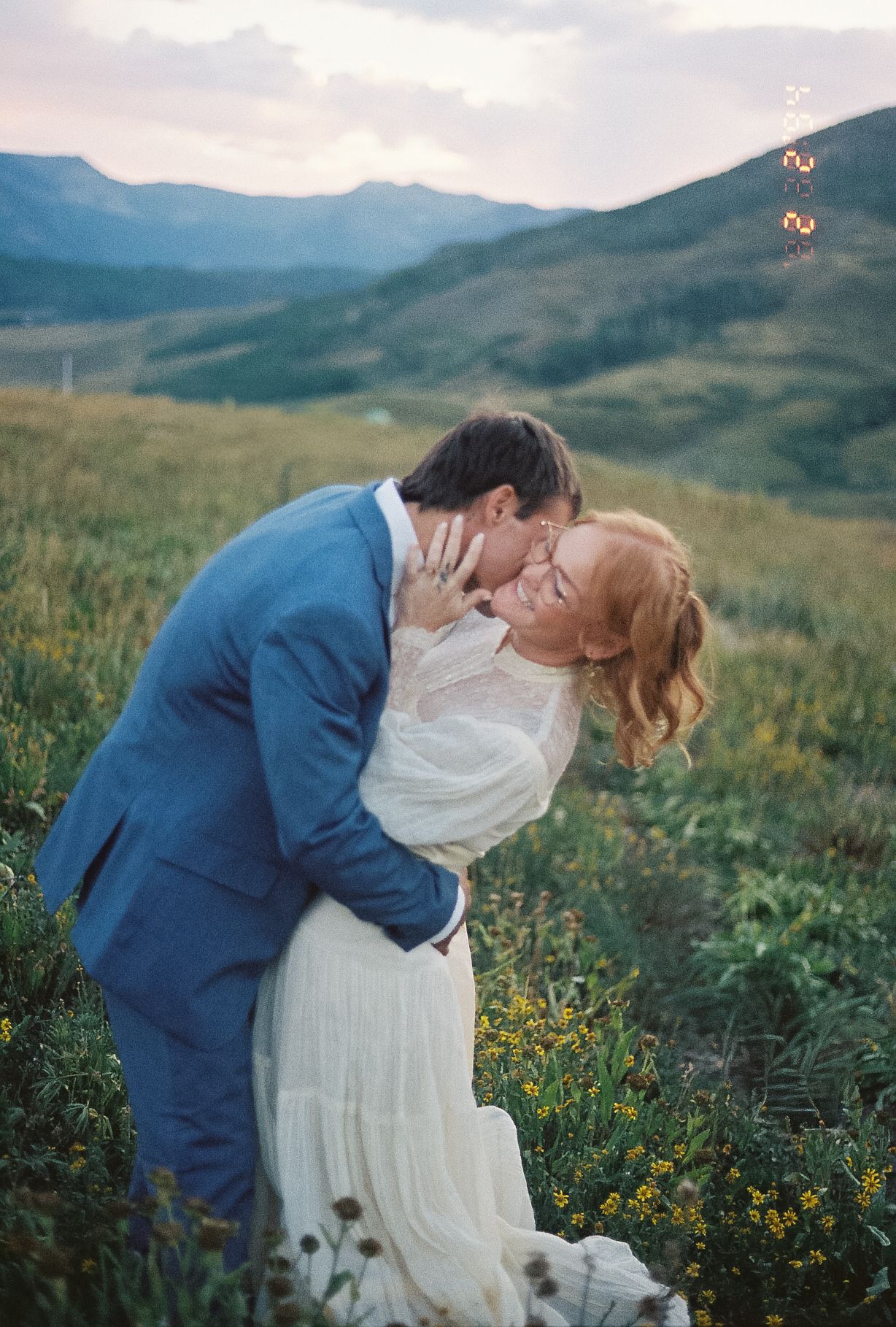 Bride and groom share romantic kiss in lush mountain meadow at sunset, groom in blue suit, bride in white lace gown.