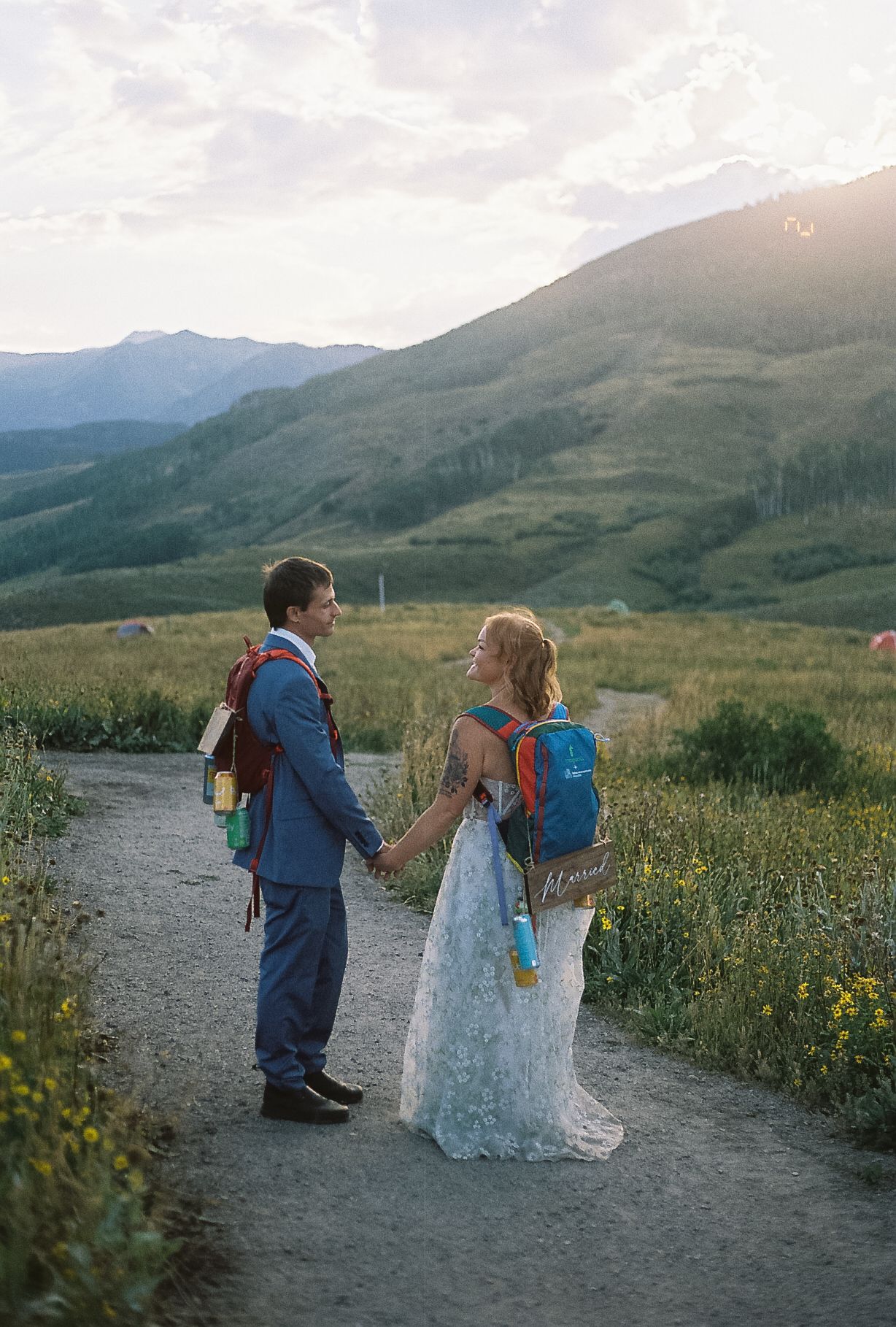Bride and groom holding hands on a trail wearing colorful backpacks with stunning mountain sunset behind them.