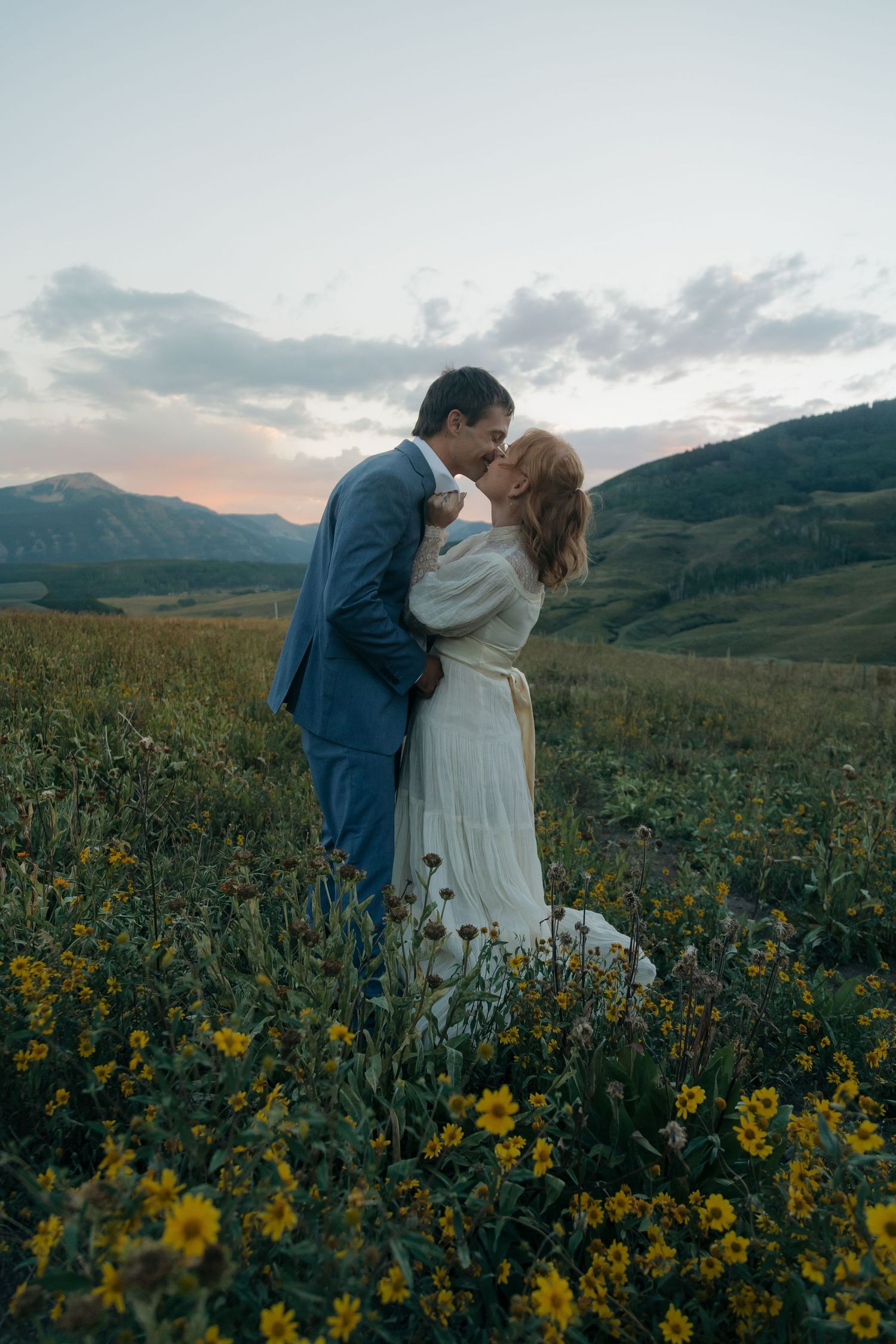 Couple shares romantic kiss in yellow wildflower field at sunset with rolling mountain landscape behind.