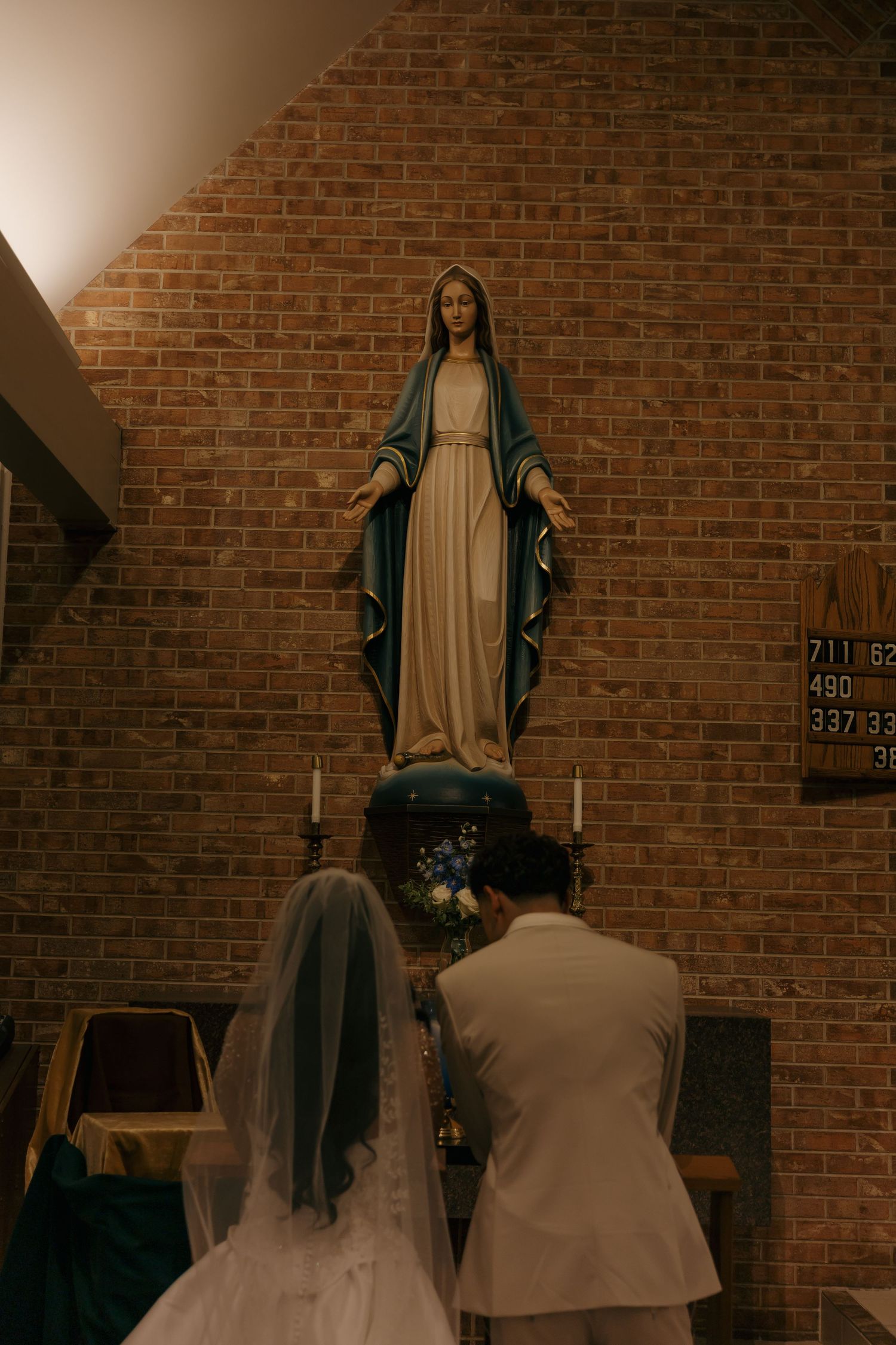 Bride and groom kneeling before Virgin Mary statue in brick church during wedding ceremony.