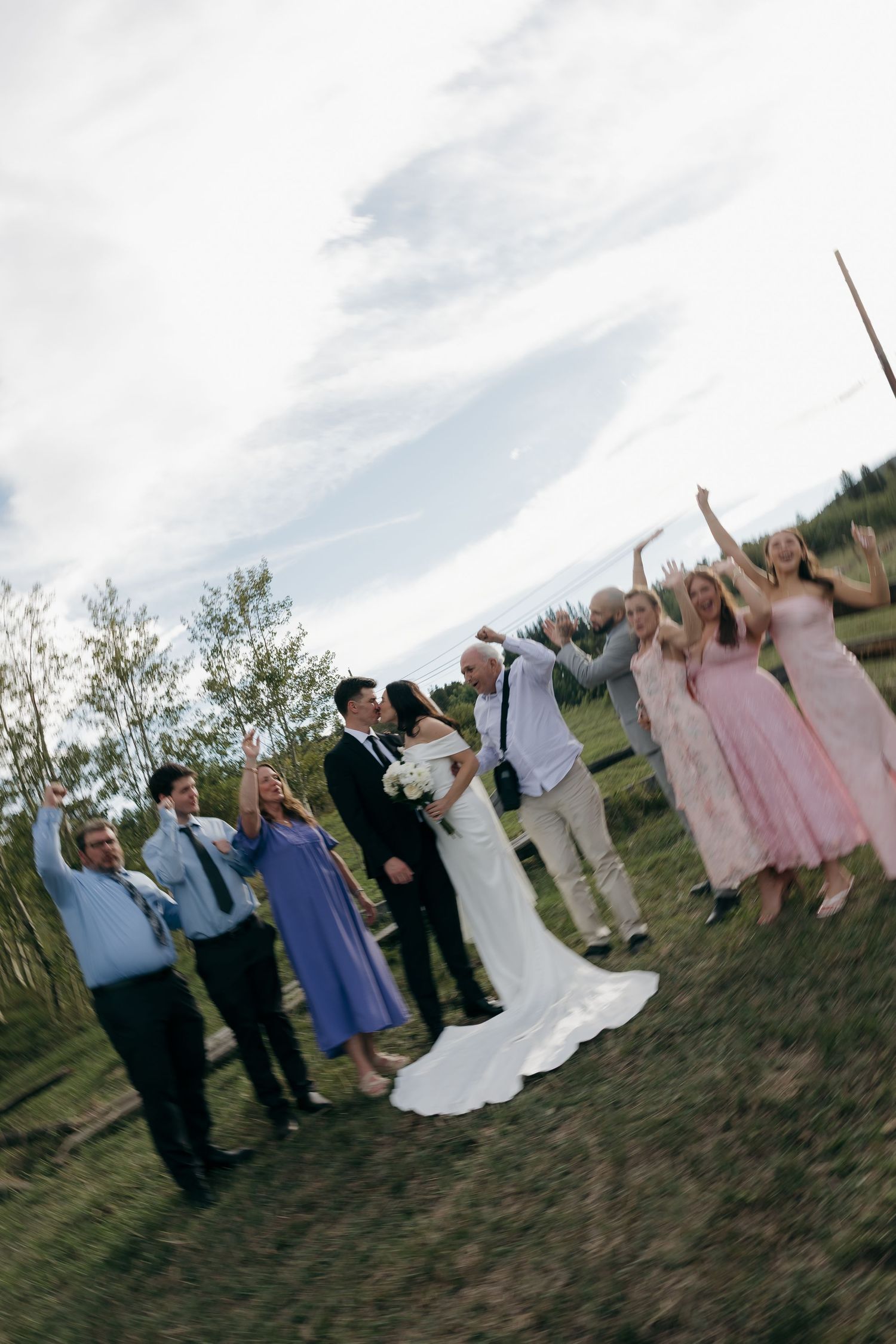 Wedding party celebrates outdoors as bride and groom kiss, guests in pink and blue attire cheer with raised arms.