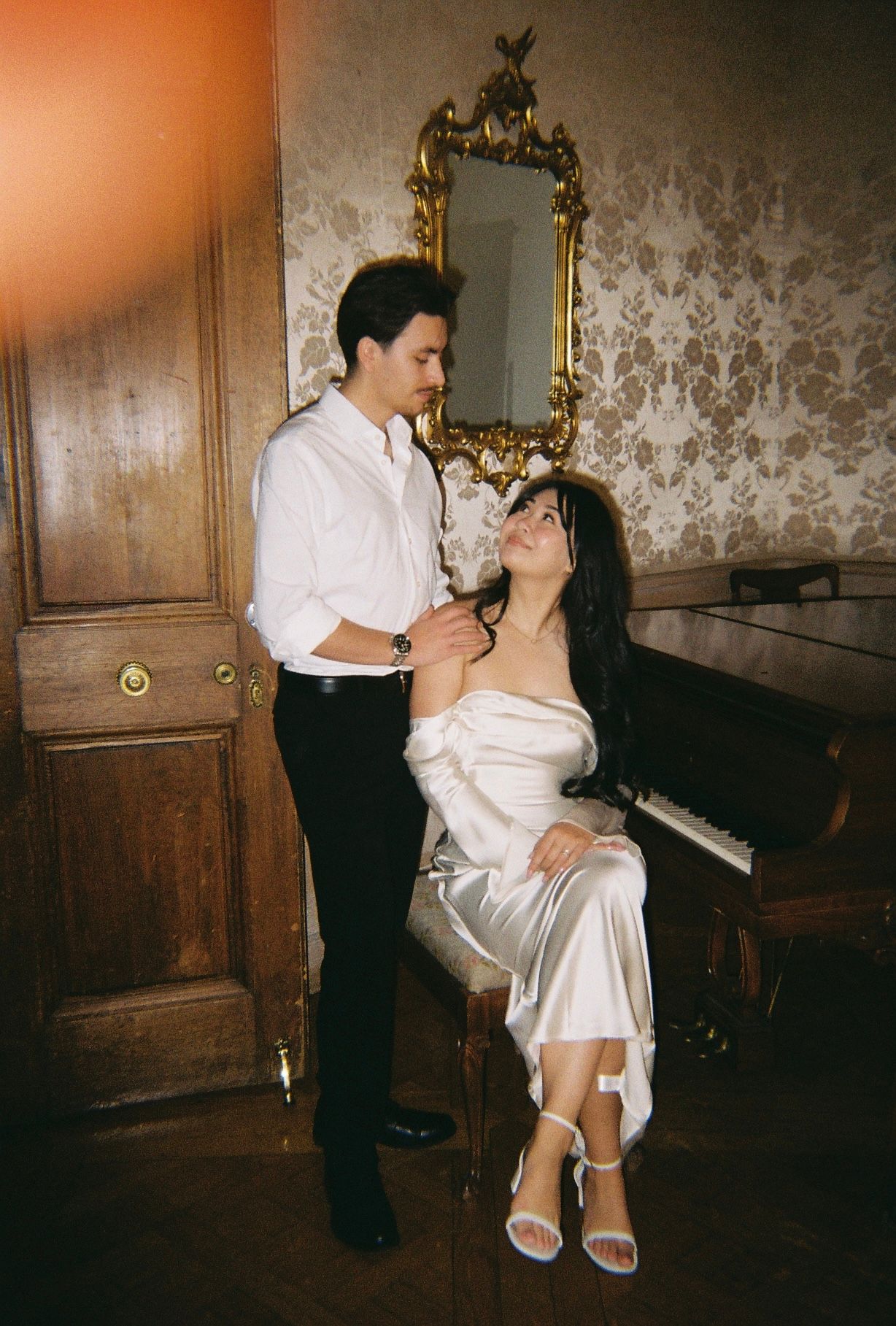 A romantic couple shares an intimate moment near a piano in a vintage-styled room with patterned wallpaper.