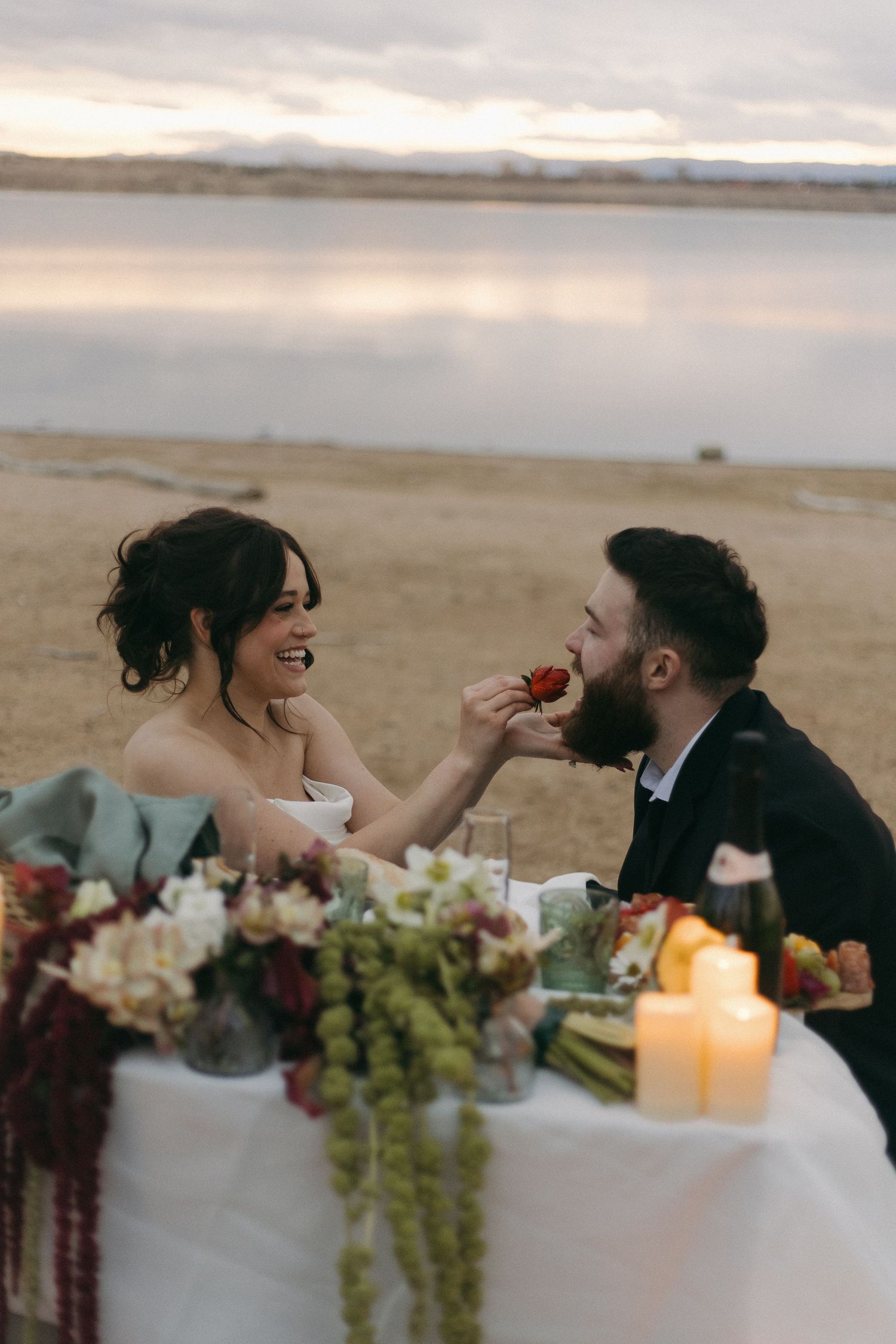 Romantic beach wedding scene with table setting featuring floral arrangements and glowing candles at sunset.