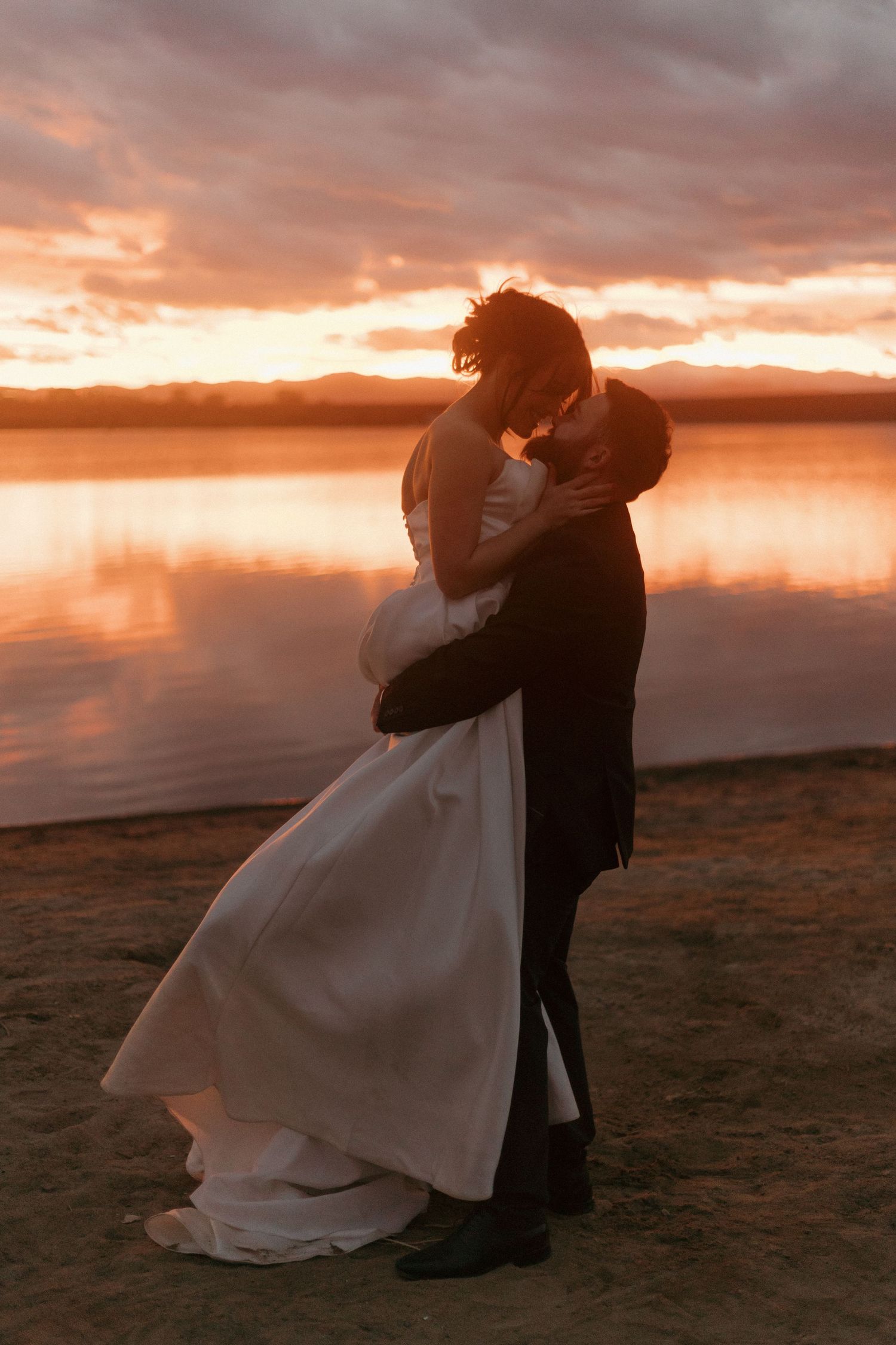 Newlyweds share a romantic moment on a beach during golden hour with mountains in the background.