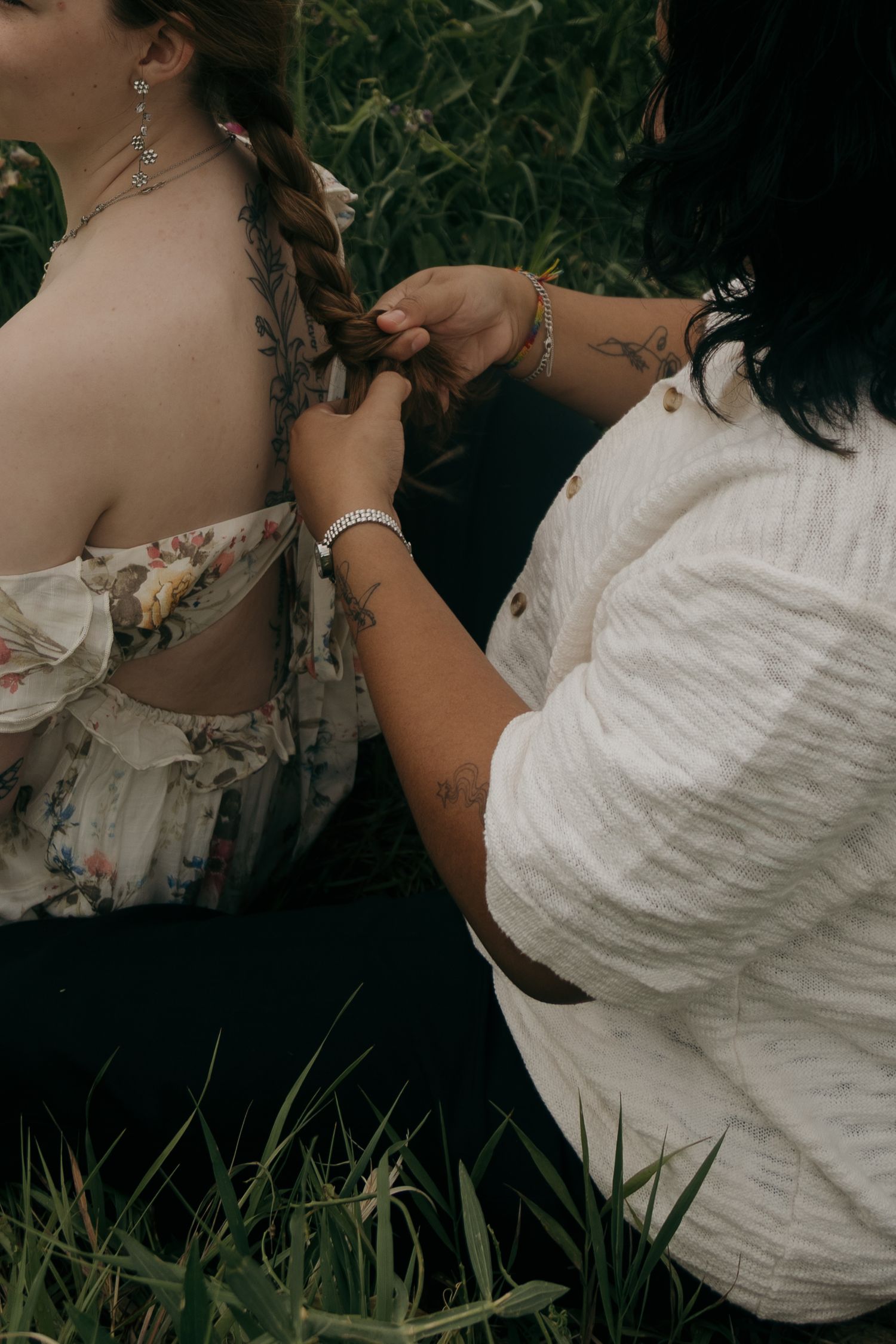 Woman braiding another woman's hair outdoors, both wearing white tops, surrounded by lush green grass.