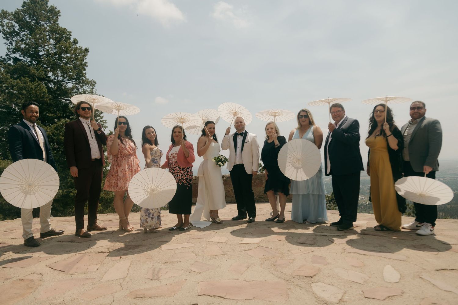 Wedding party group photo on mountain overlook holding white parasols under blue sky.