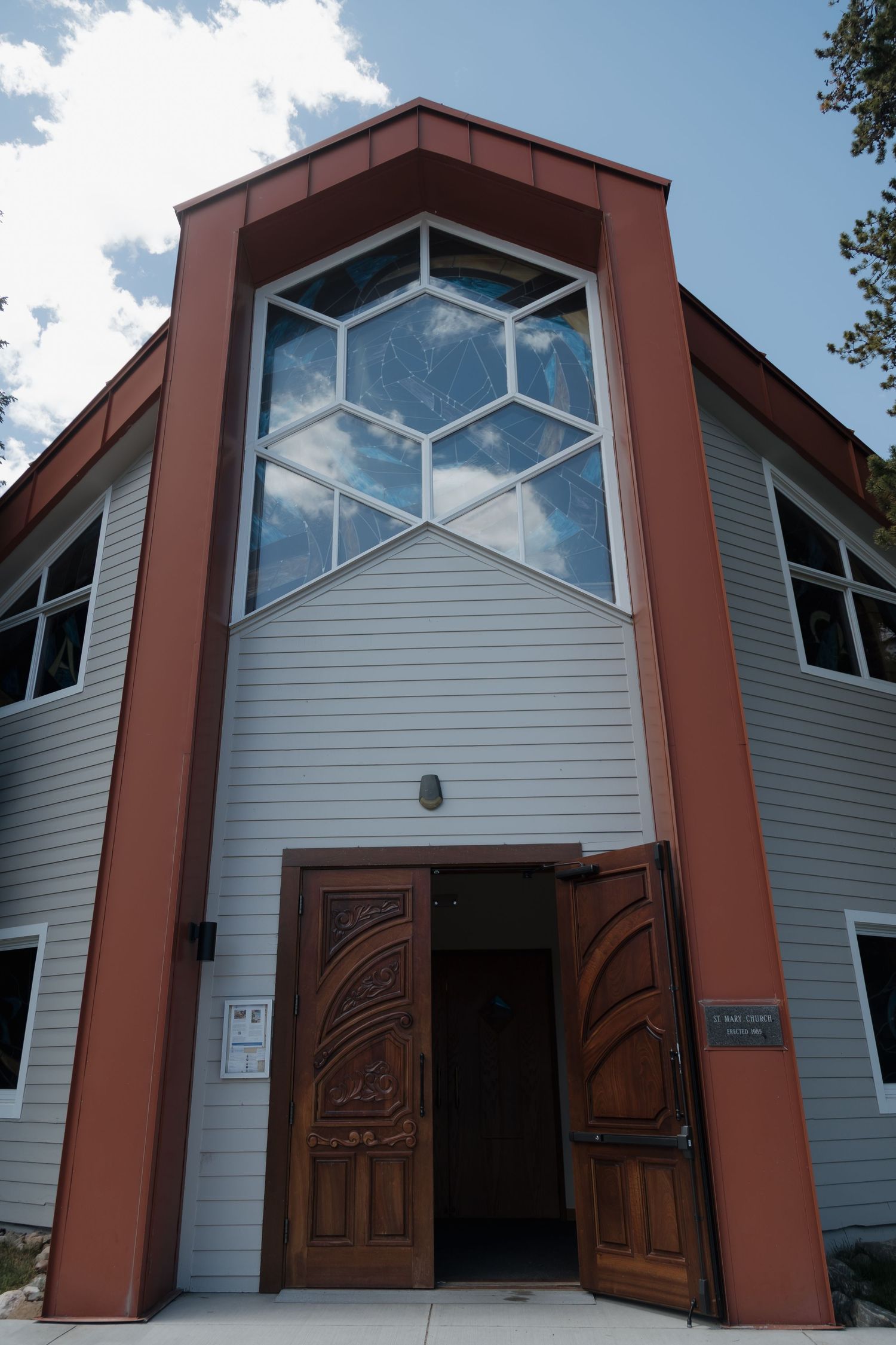 Modern church entrance with large hexagonal stained glass window, ornate wooden double doors, and red trim facade.