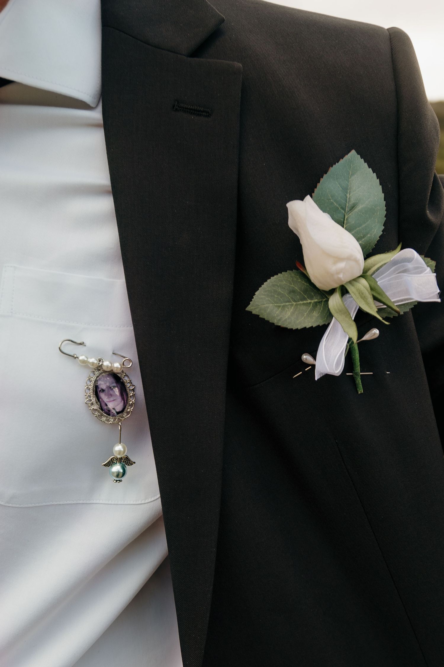 Groom's black tuxedo lapel with white rose boutonniere and vintage brooch memorial pin on white dress shirt.