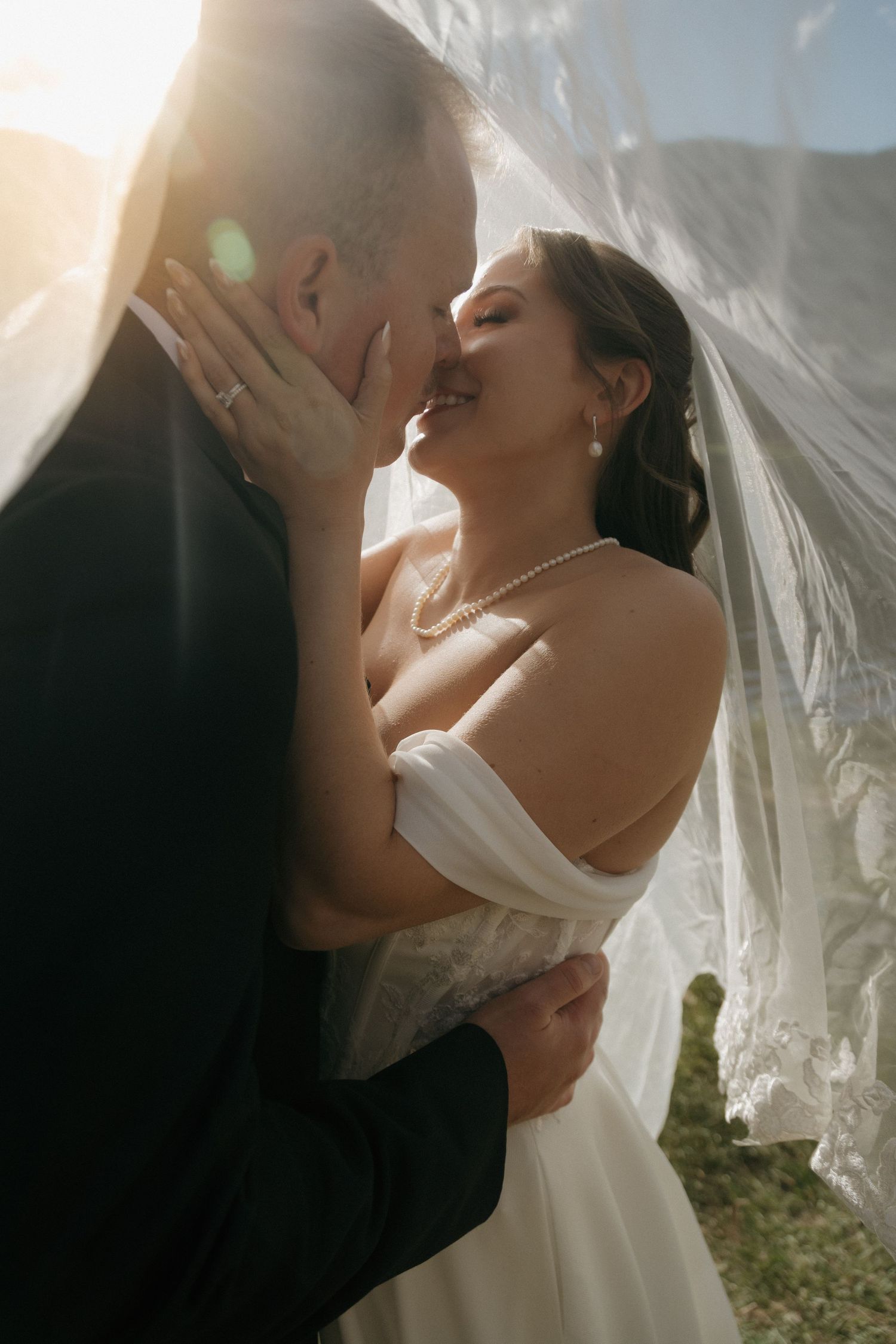 Bride and groom share an intimate moment under a flowing veil with stunning golden sunlight backlighting the scene.