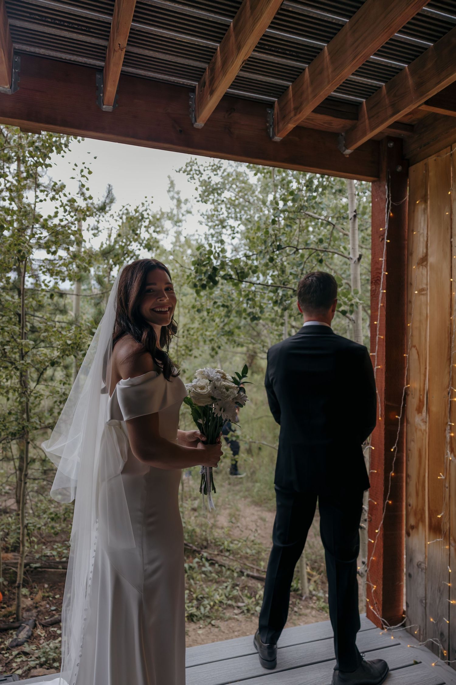 Bride in white off-shoulder gown holding bouquet laughs during first look moment on rustic wooden deck.