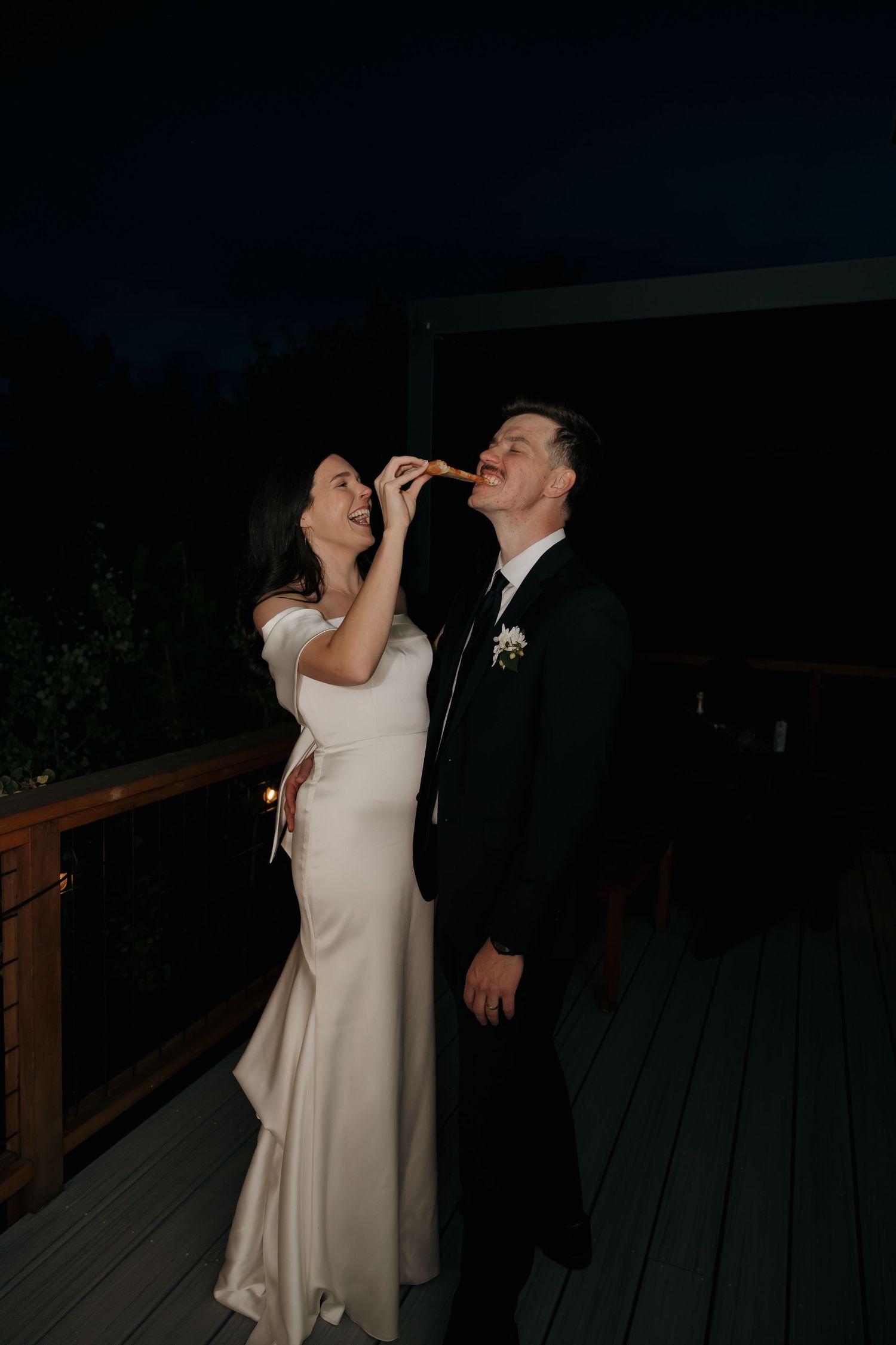 Bride feeds groom a slice of pizza on dark outdoor deck during wedding reception at night.