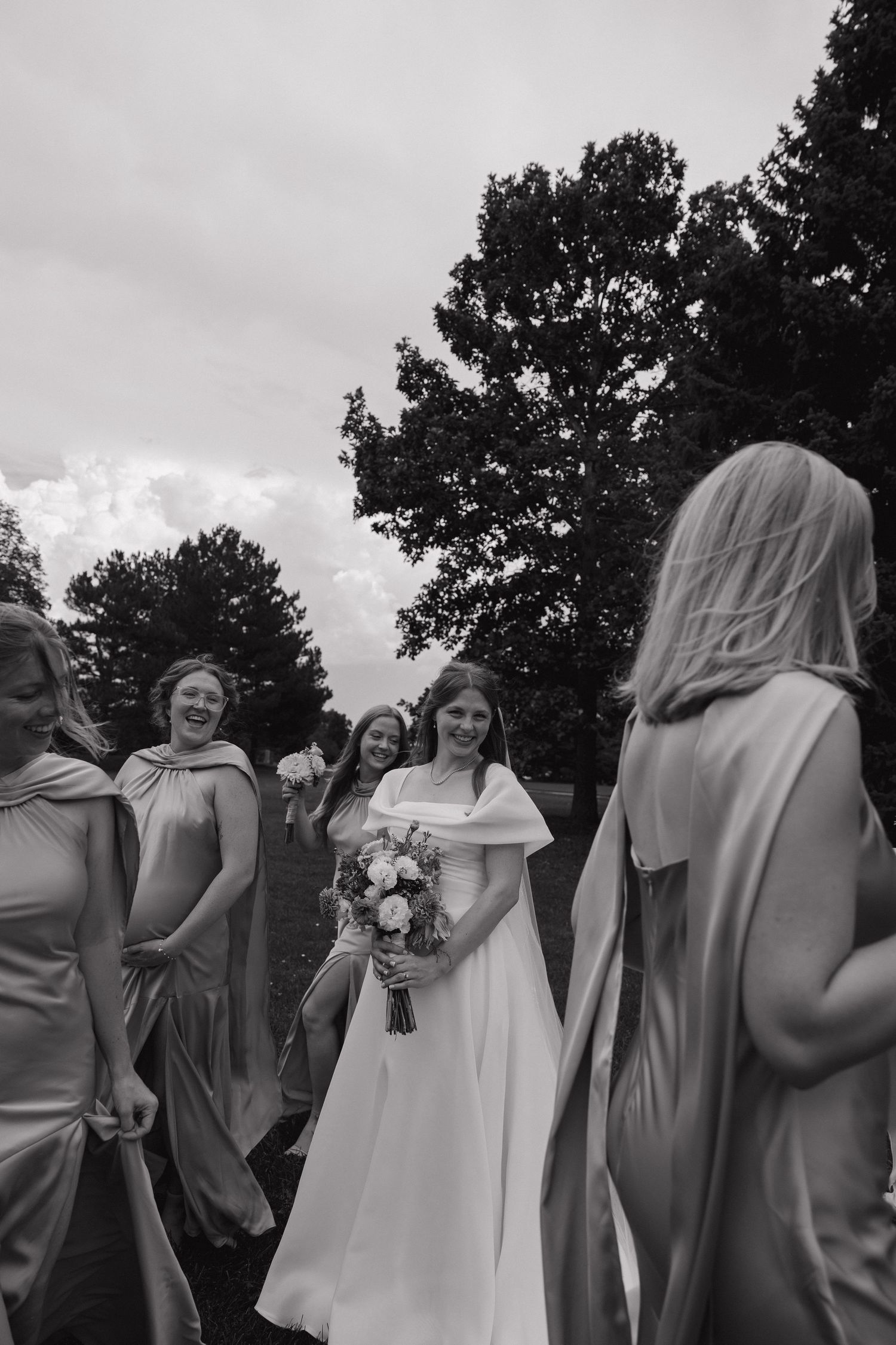 Bride in white gown holds bouquet surrounded by smiling bridesmaids in satin dresses outdoors in black and white photo.