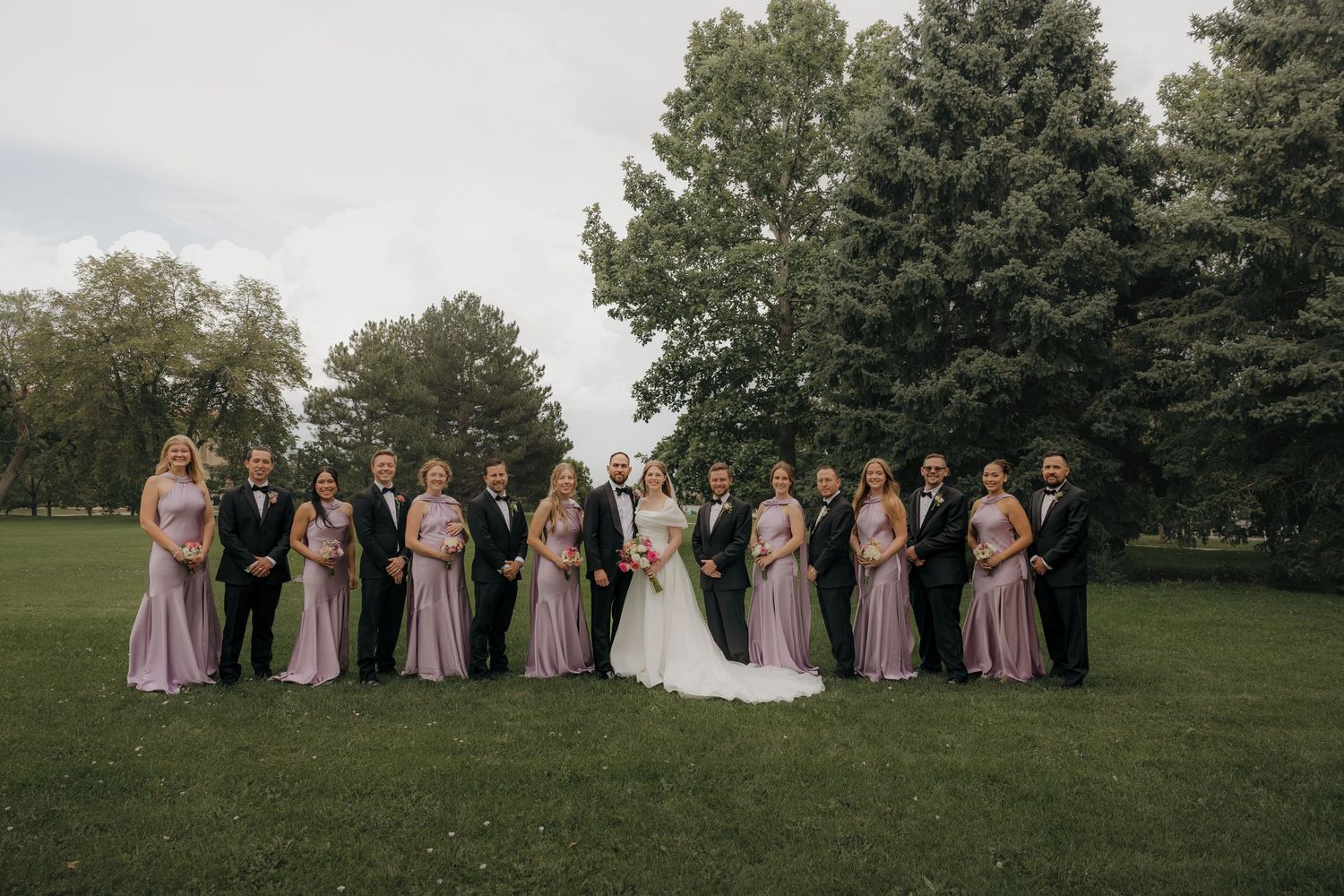 Wedding party poses outdoors with bridesmaids in lavender dresses on green lawn.