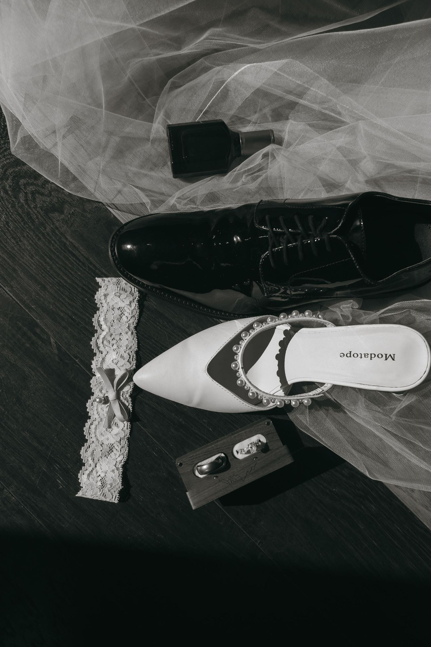 Black and white flat lay of wedding accessories including bridal heels, groom shoes, veil, garter, rings, and cologne.