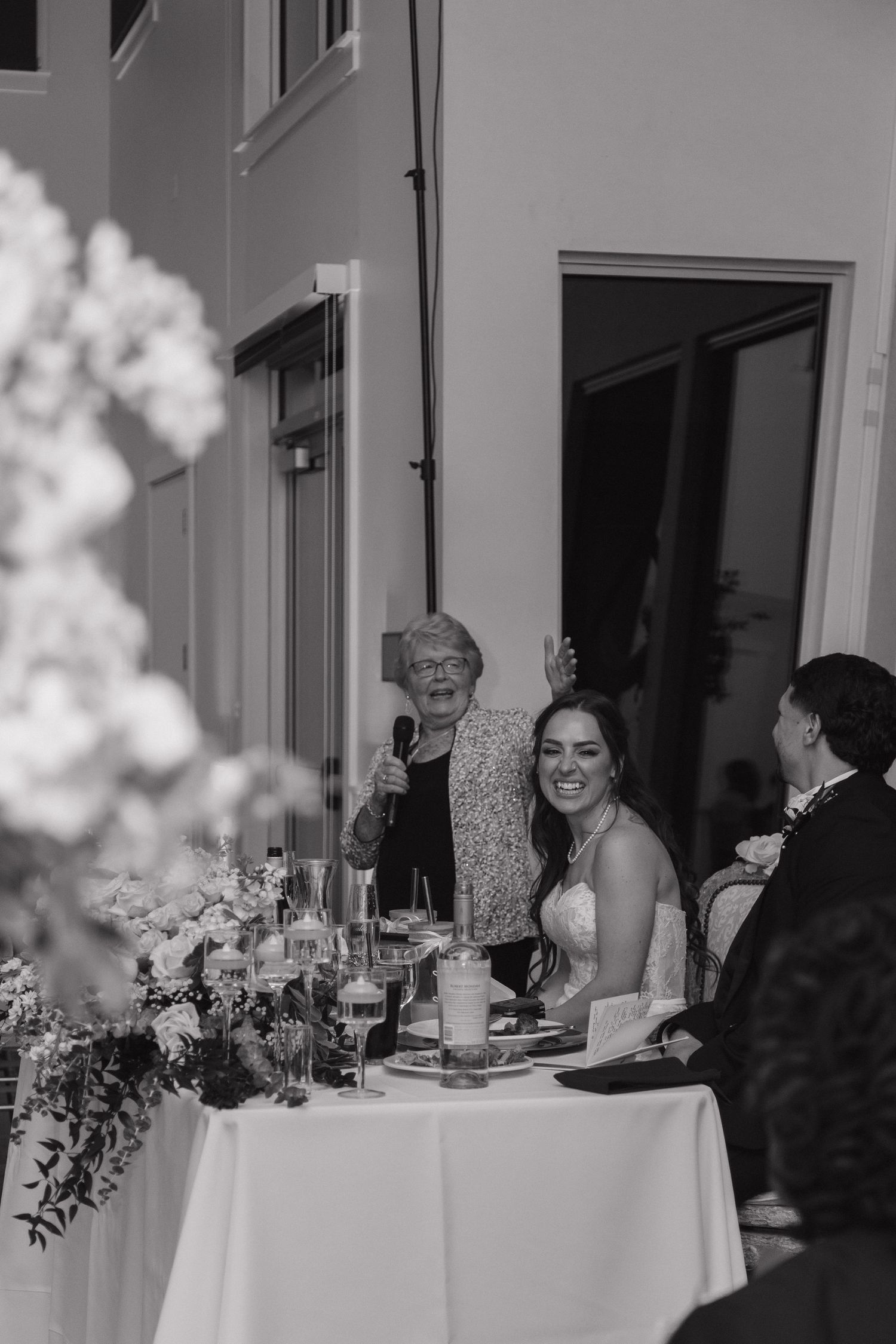 Bride smiles as an older woman gives a heartfelt speech at the wedding reception head table in black and white.