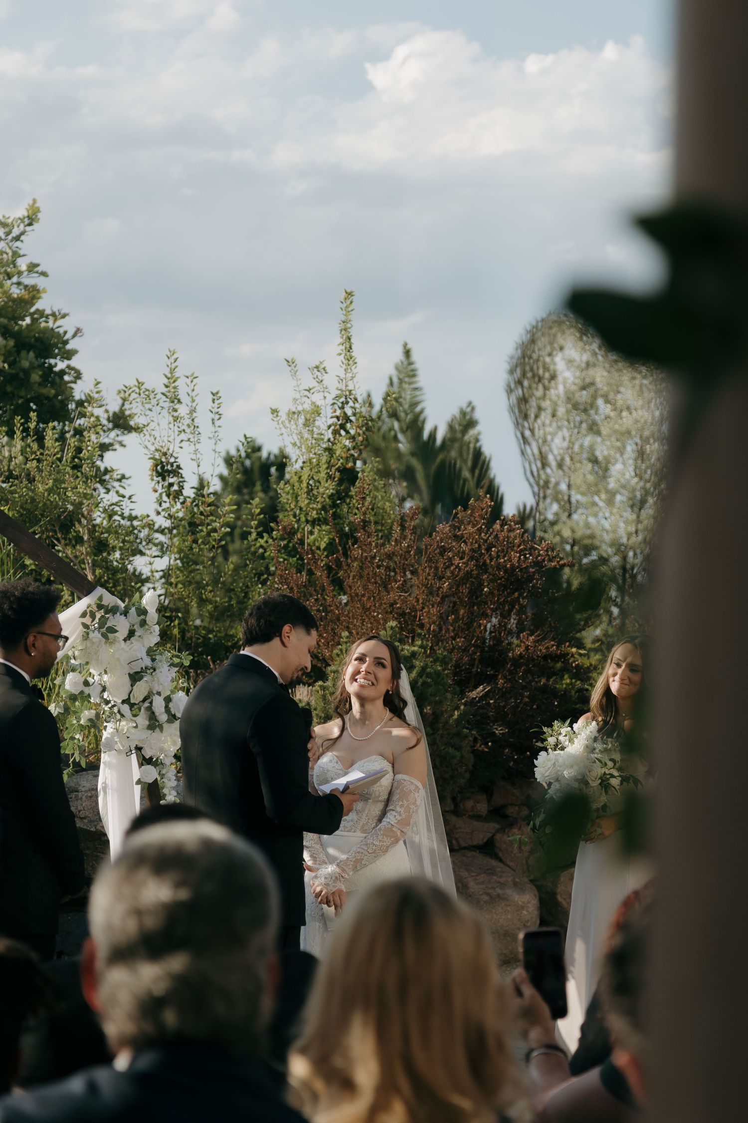 Radiant bride smiles at groom during outdoor wedding ceremony surrounded by lush greenery.