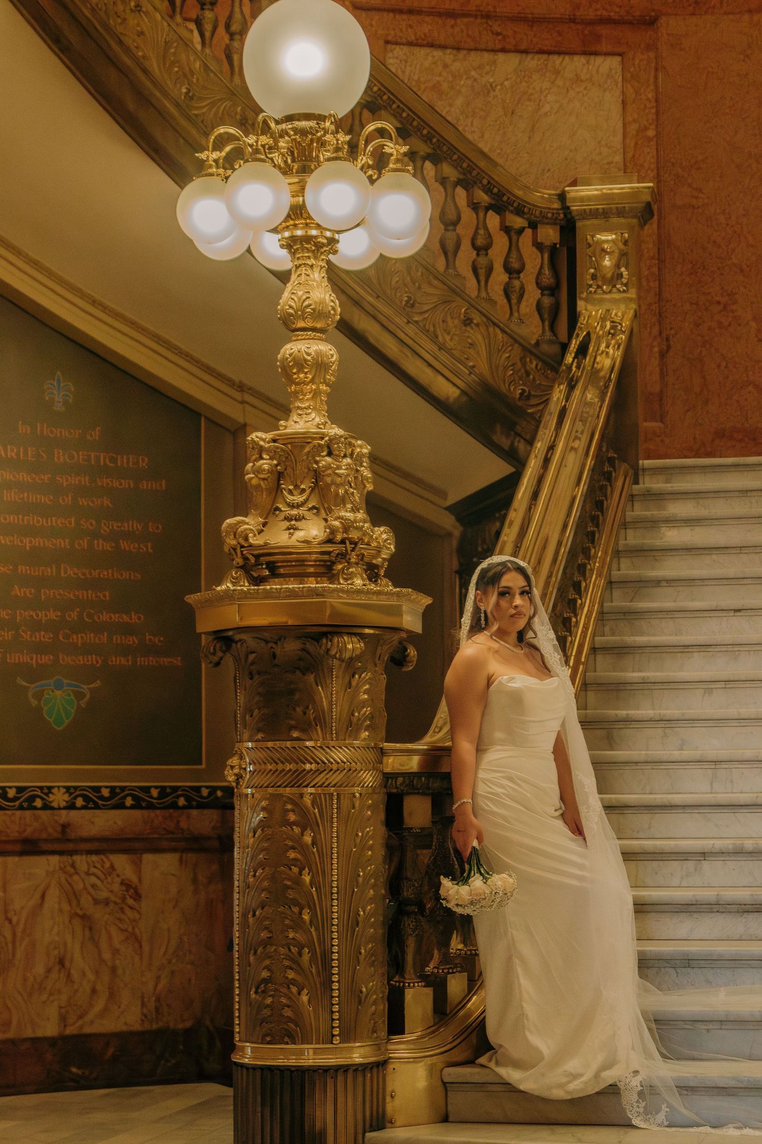 Elegant white gown photographed against ornate gold-detailed staircase and vintage light fixture in historic building interior.