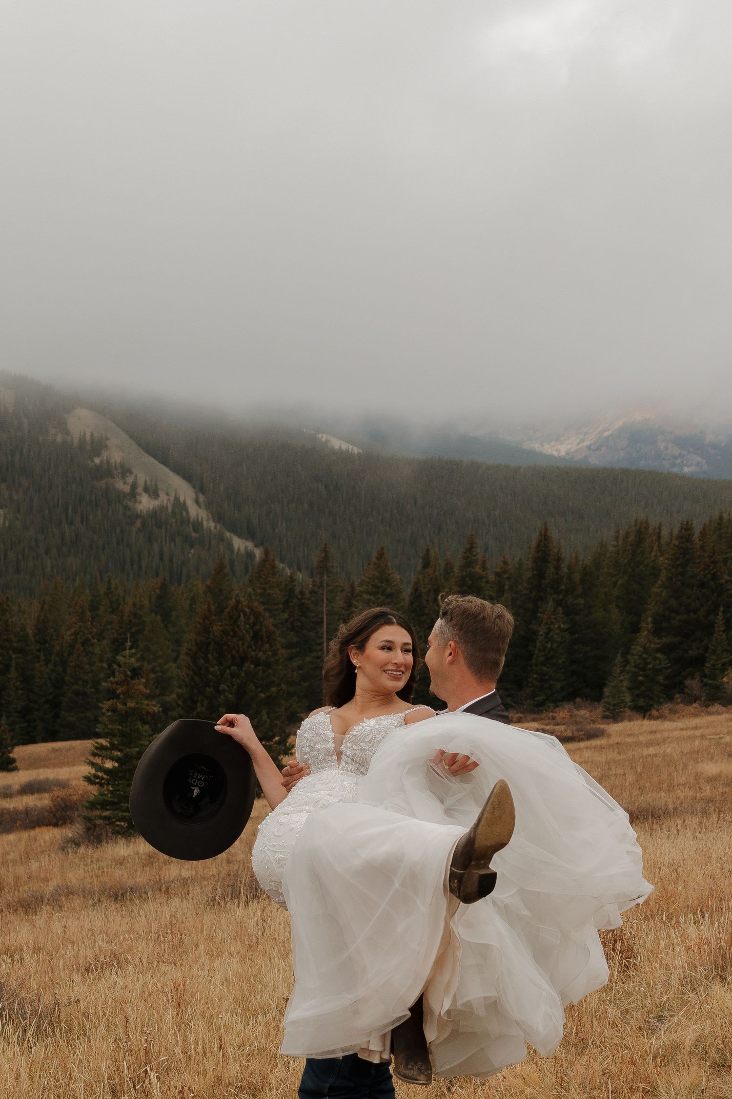 Newlyweds dance and twirl playfully in a mountain meadow with evergreen forests behind them.