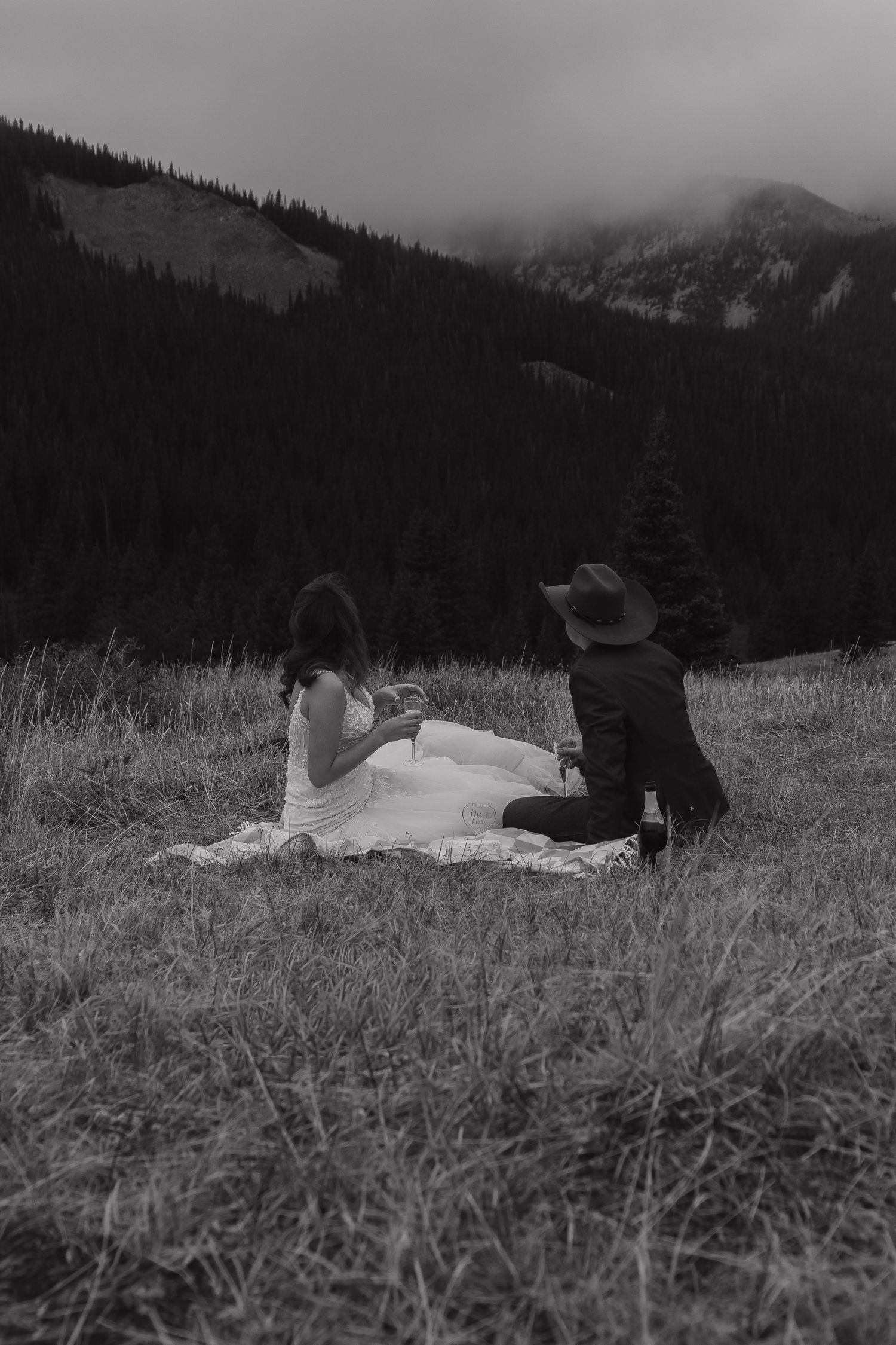 A black and white photo of a couple lying in tall mountain grass with evergreen forests behind them.