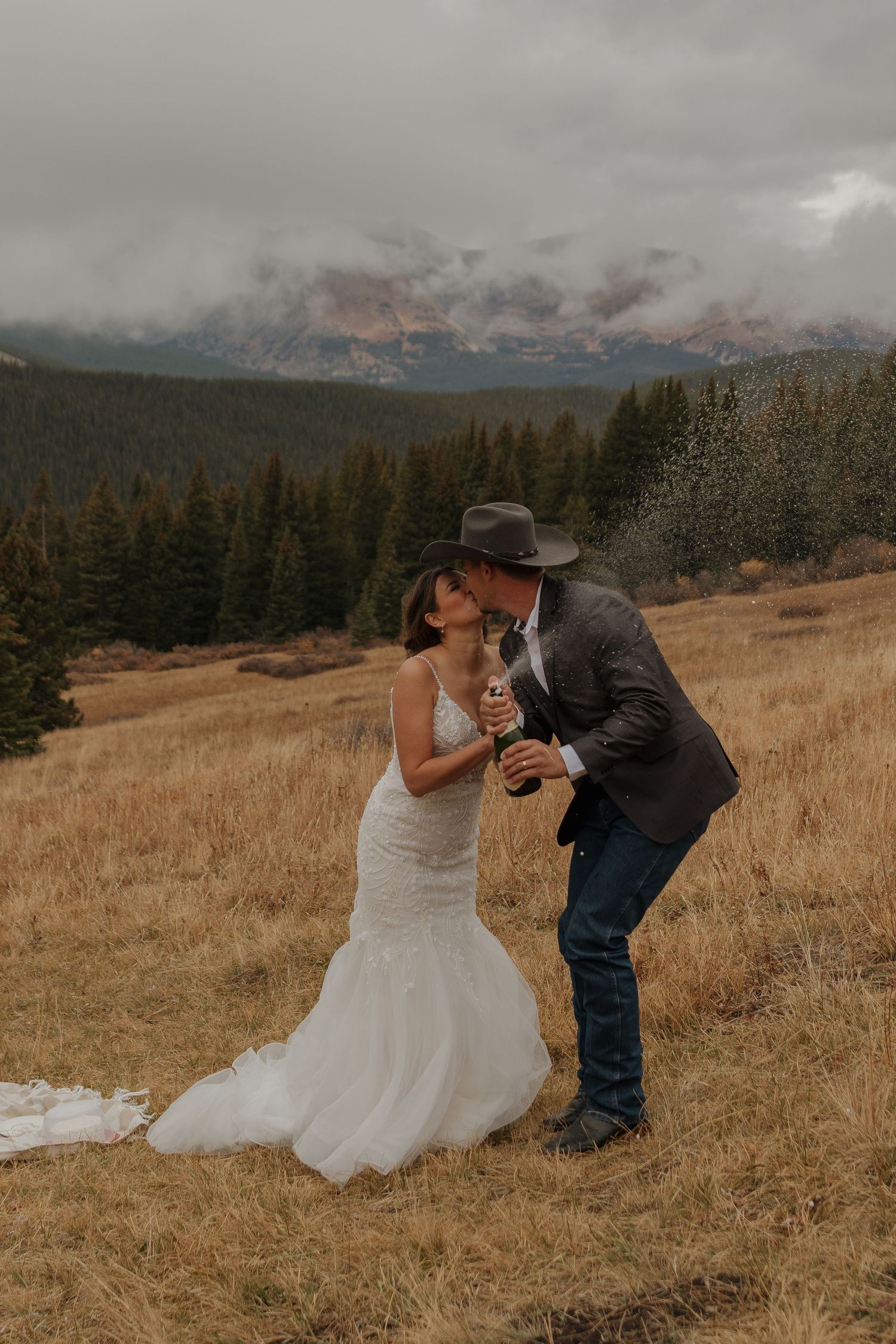 A couple shares a playful moment in a mountain meadow with dramatic cloudy peaks in background.