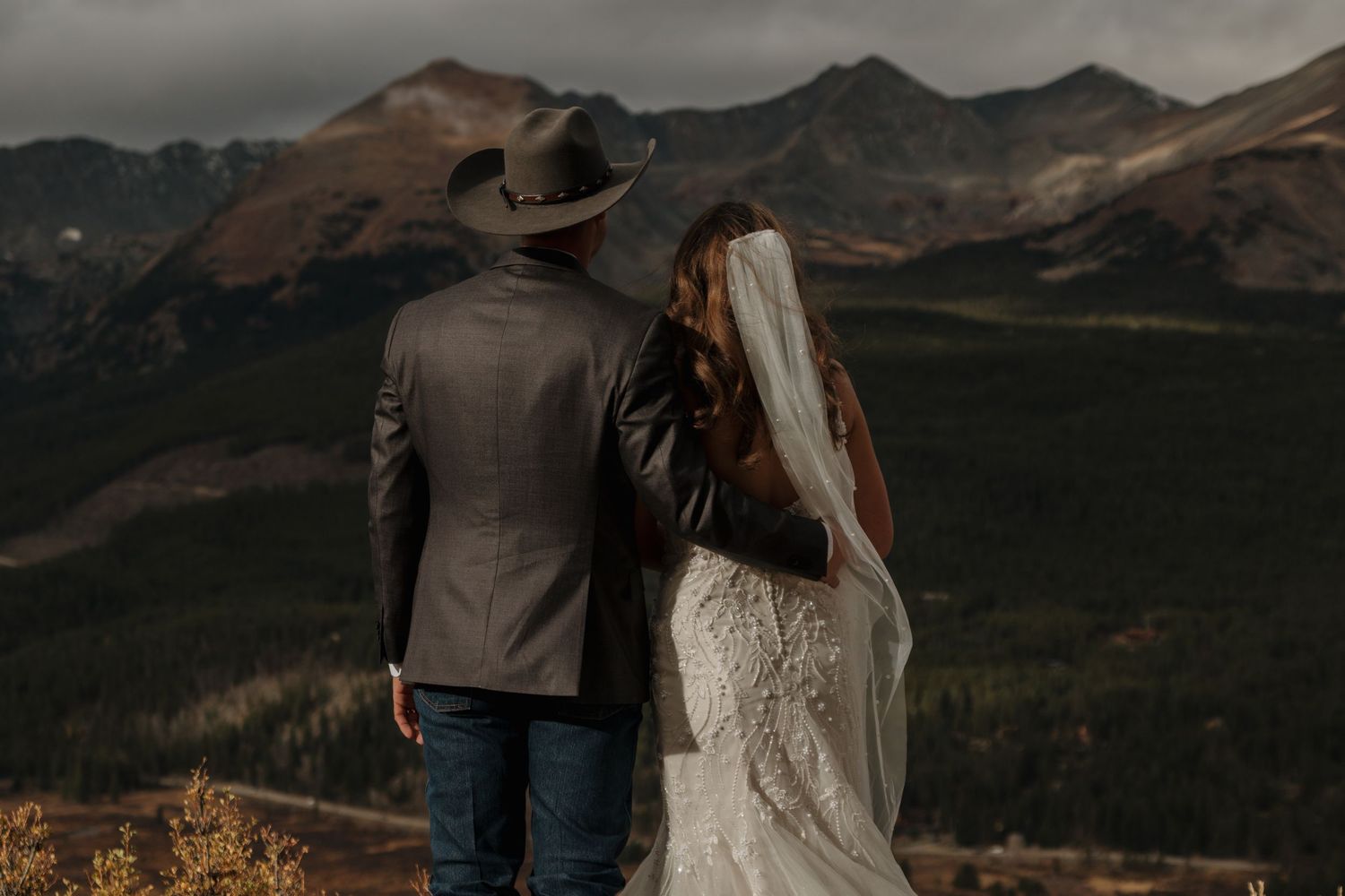 A couple in western attire stands together overlooking a dramatic mountain landscape.