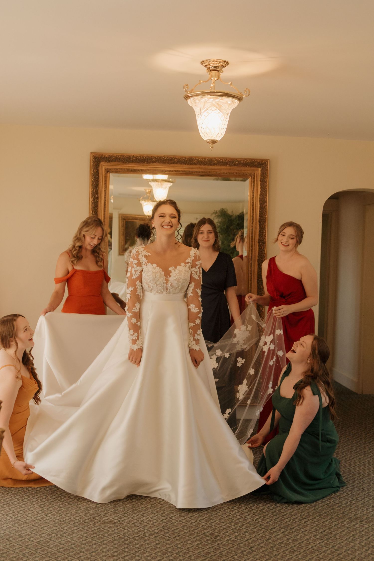 A bride in a lace-sleeve wedding gown stands surrounded by attendants in colorful dresses.