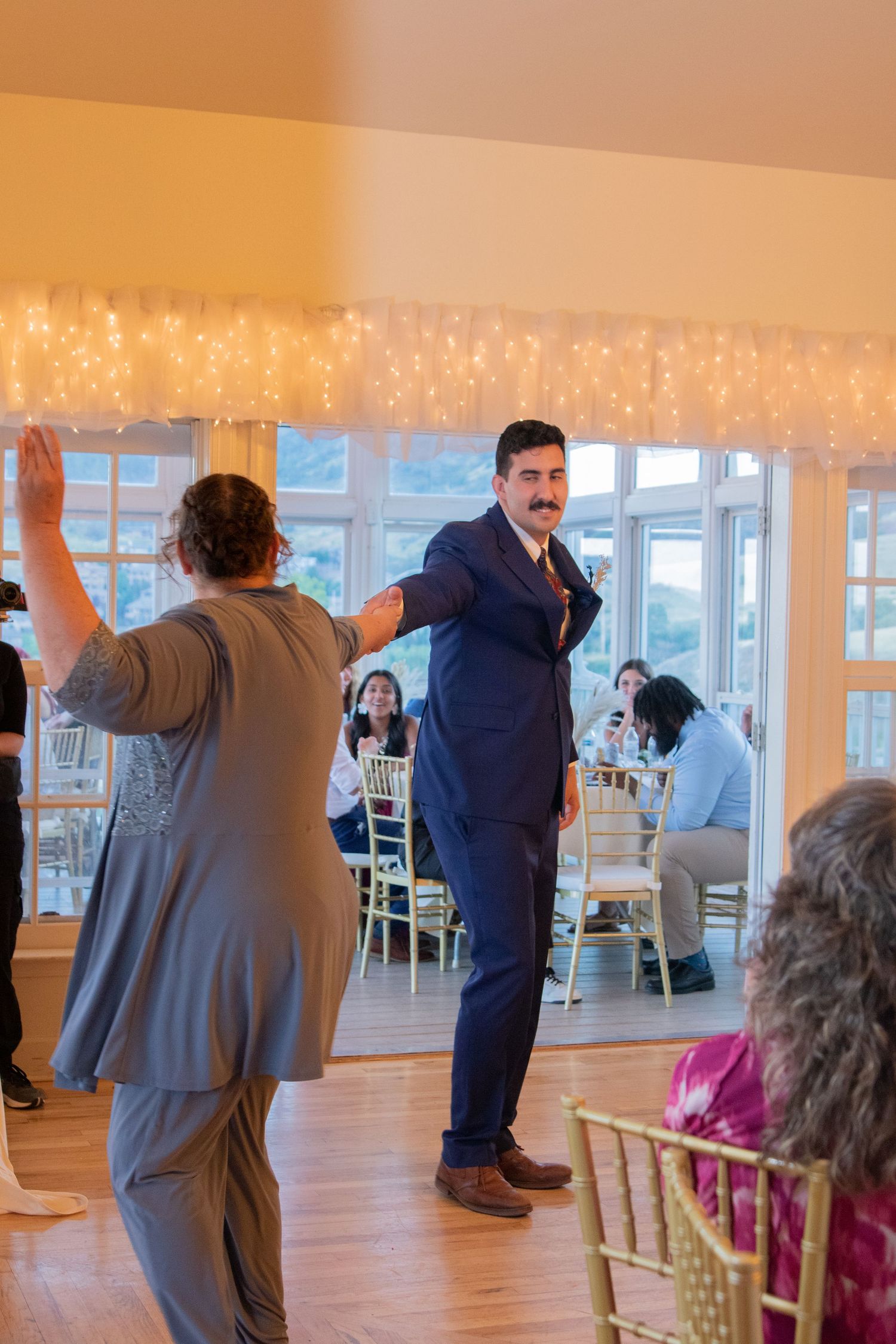 Wedding reception scene with twinkle lights and decorations while guests dance on hardwood floor.