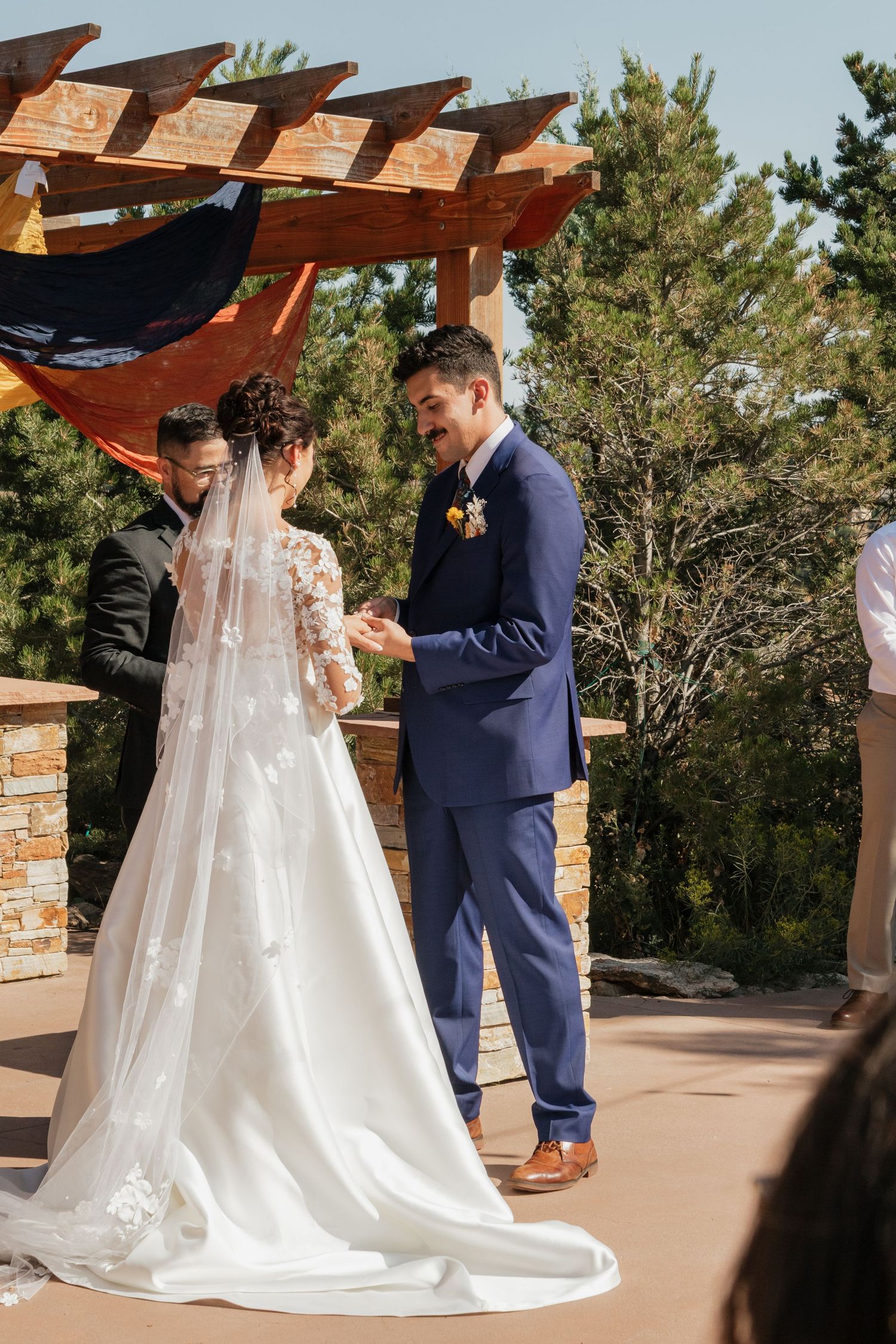 A bride in a flowing white gown and groom in navy suit share an intimate moment under a wooden pergola.