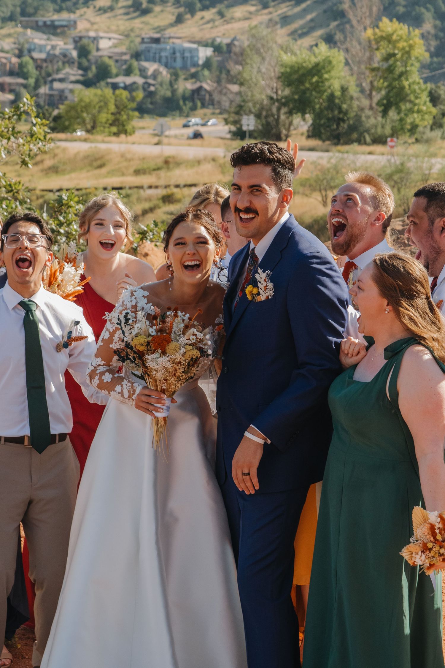 Wedding party laughs joyfully together outdoors with bridesmaids in green dresses and autumn-colored bouquets.