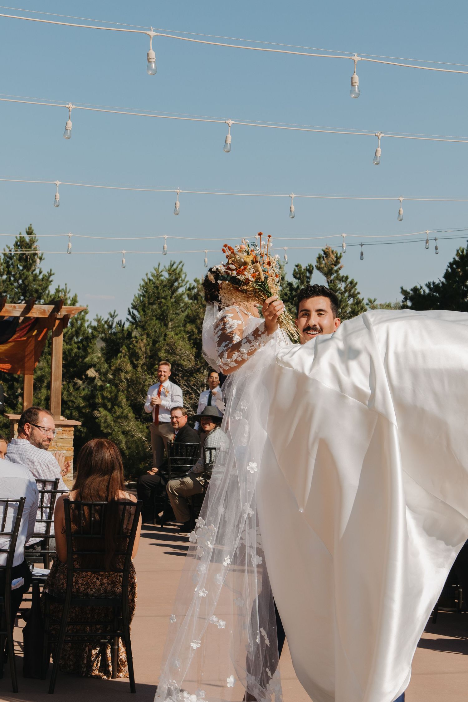 String lights hang above an outdoor wedding venue against a bright blue sky.