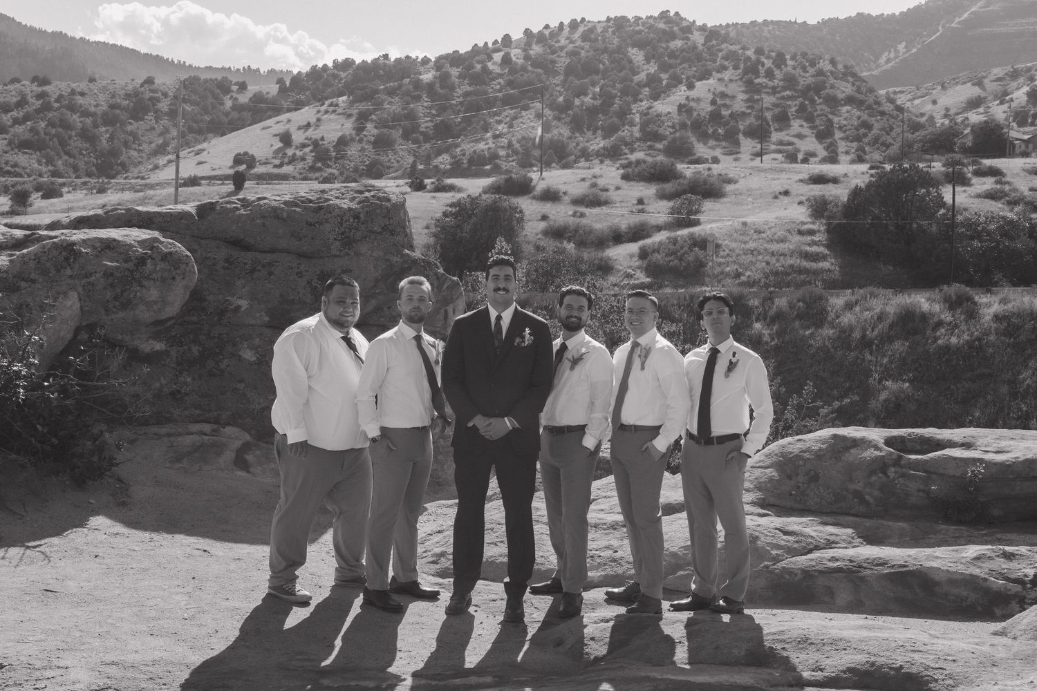 Wedding party in neutral colors poses on rocky terrain with mountains in background.