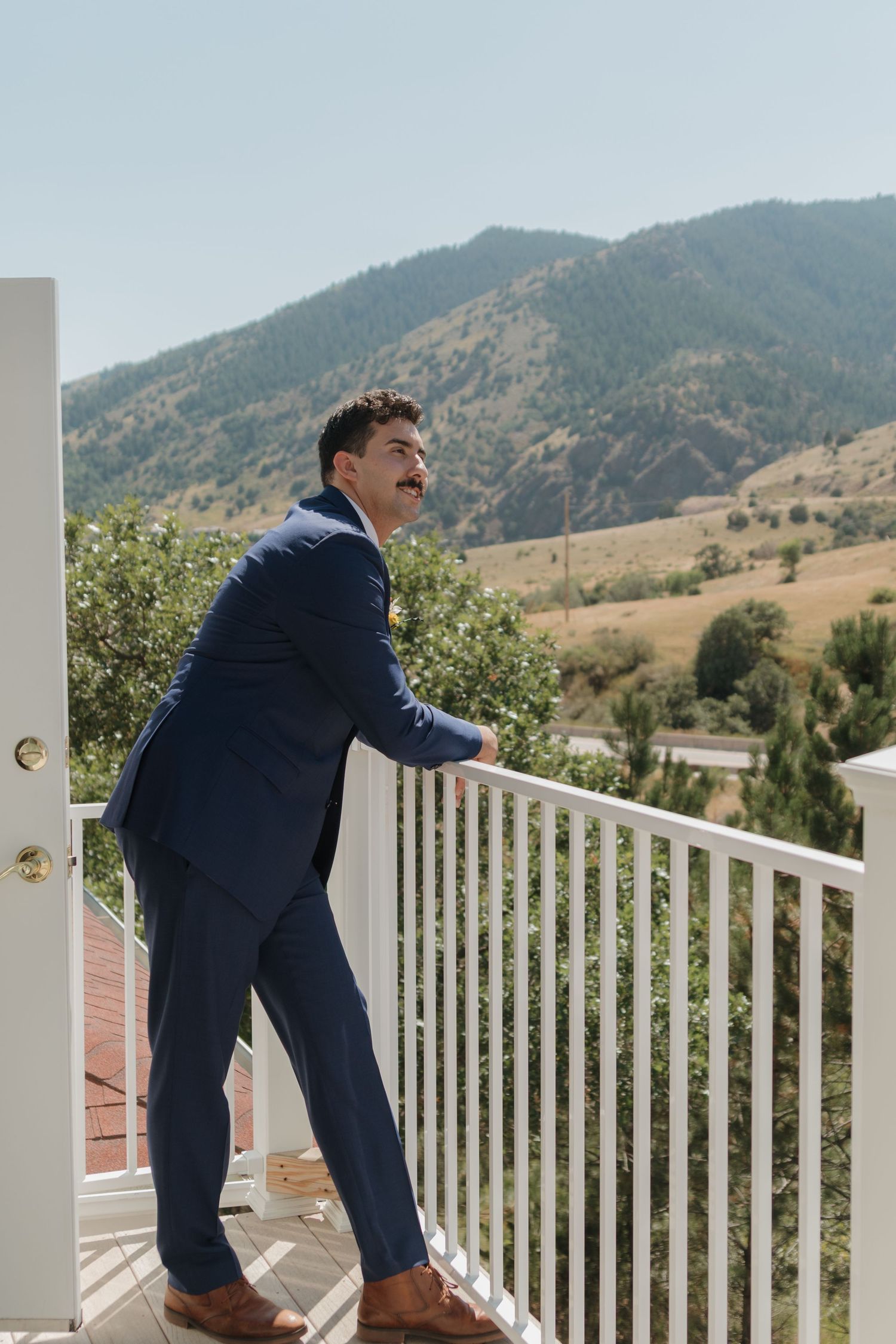 Groom in navy suit climbs over white porch railing with mountains in background.
