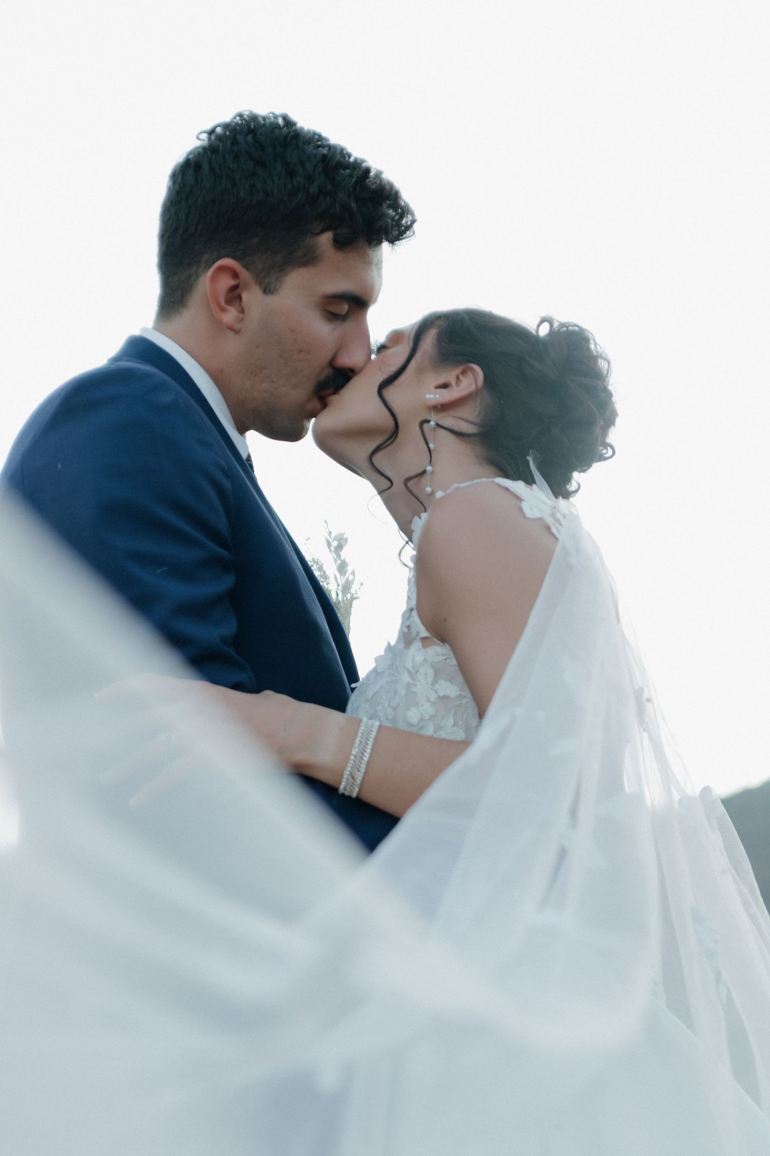 Romantic black and white wedding portrait with flowing veil against bright sky.