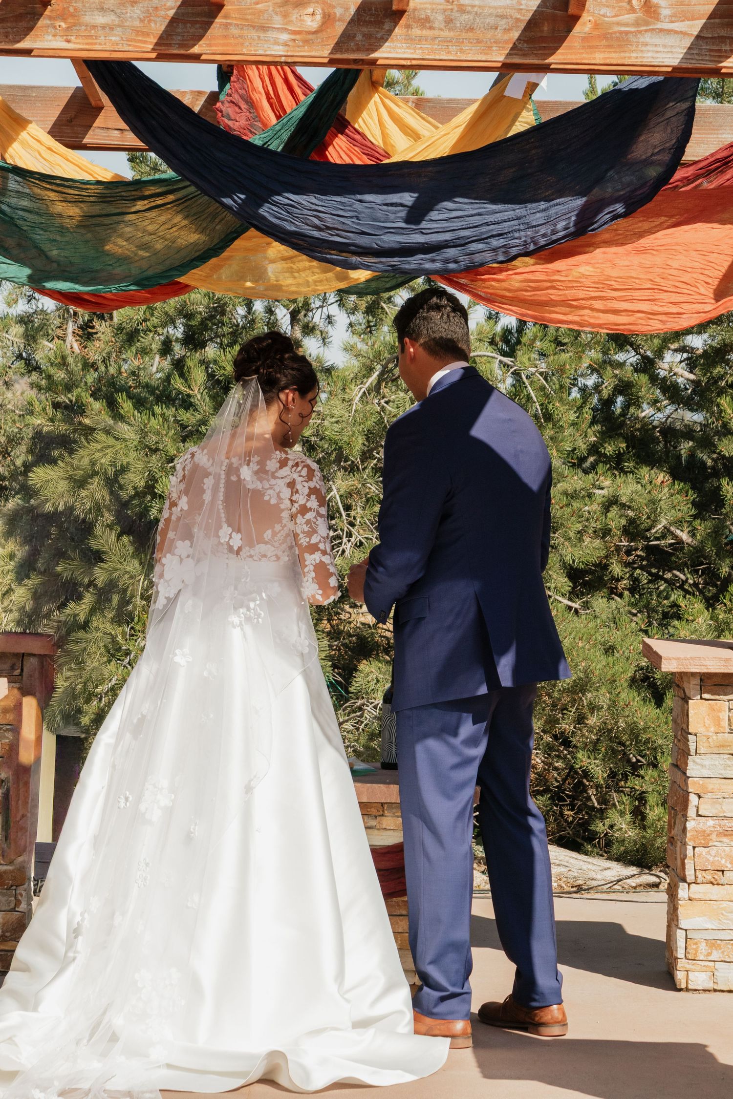 Couple stands under wooden beams during outdoor wedding ceremony with flowing white dress visible.