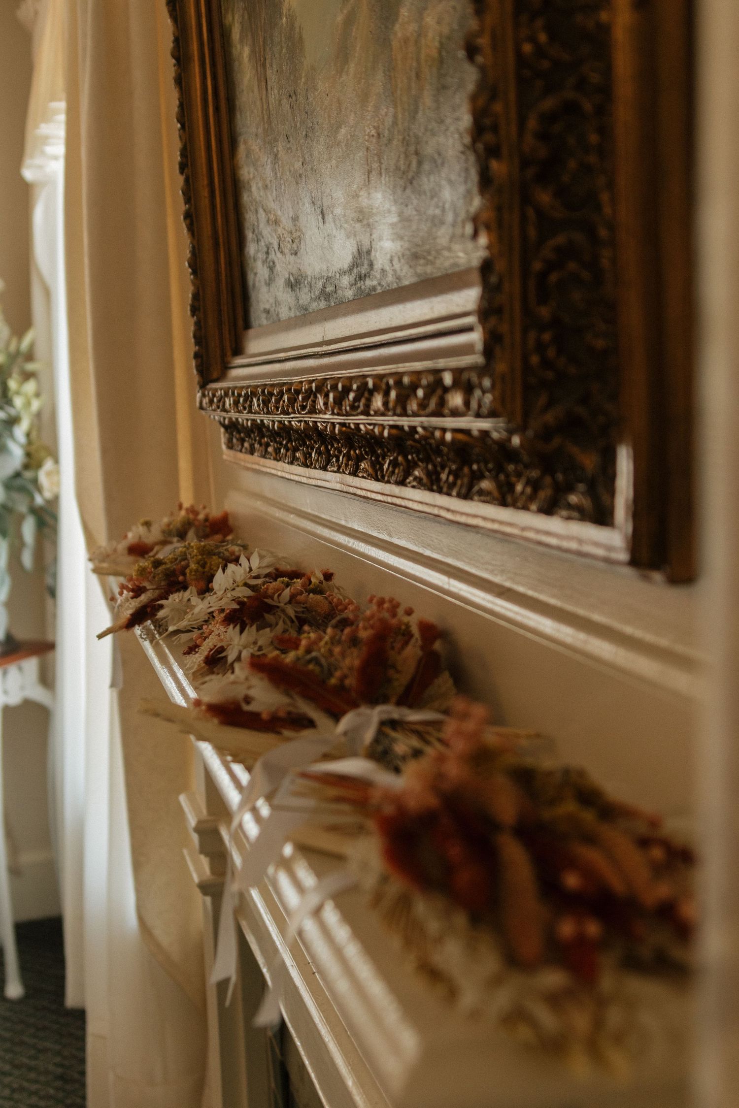 Ornate antique mirror with intricate wooden frame hangs on beige wall with dried floral arrangements beneath.
