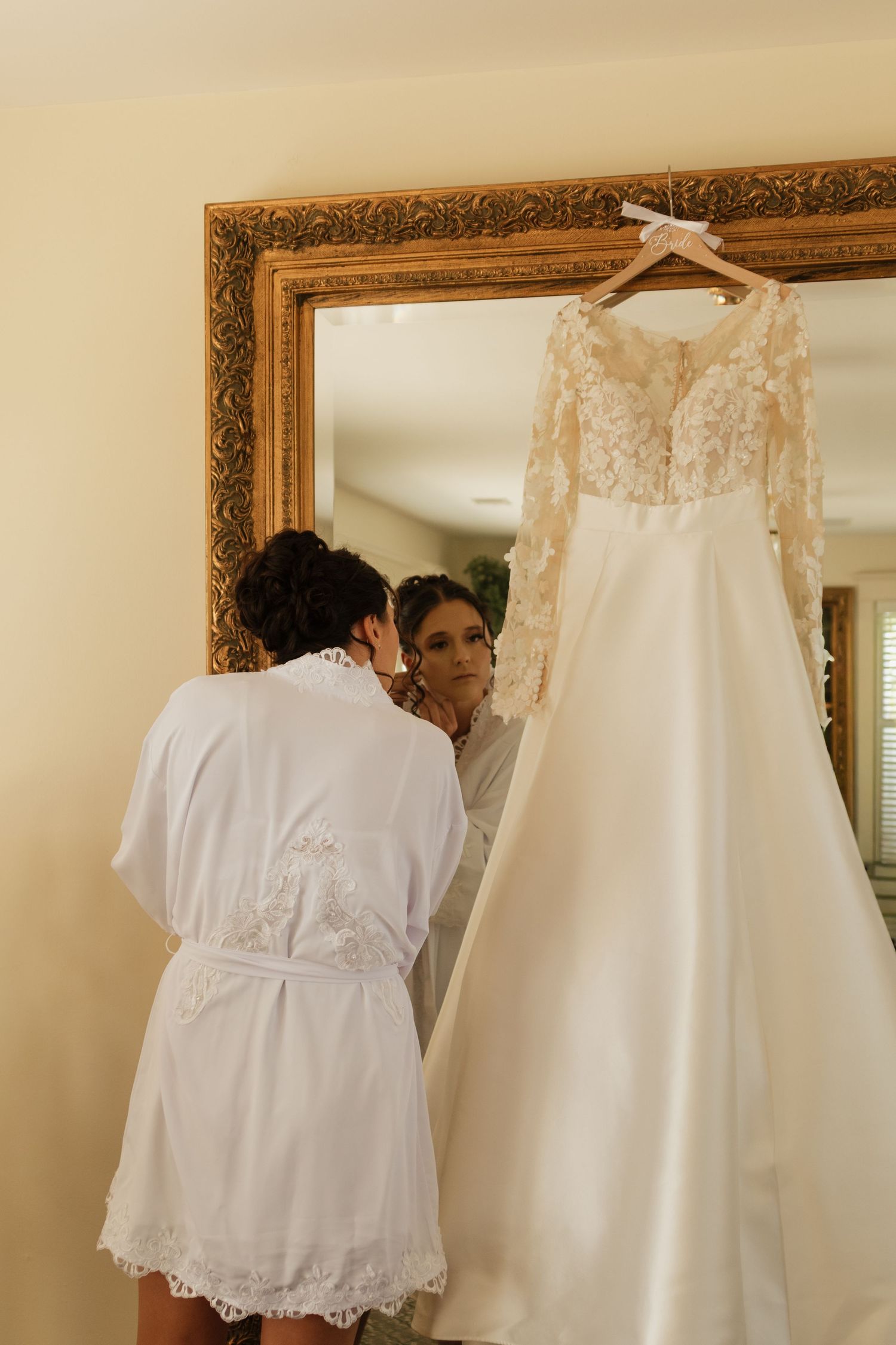 White lace dresses and gowns displayed on hangers against a wall with ornate gold-framed mirrors.