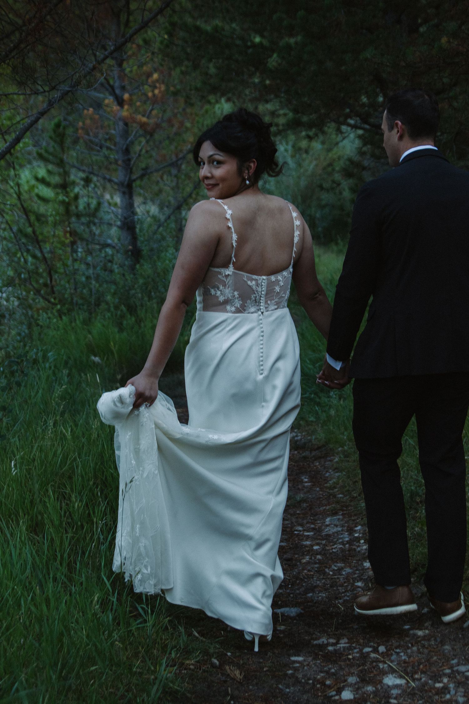Two people walk along a forest path while holding a flowing white wedding dress train.