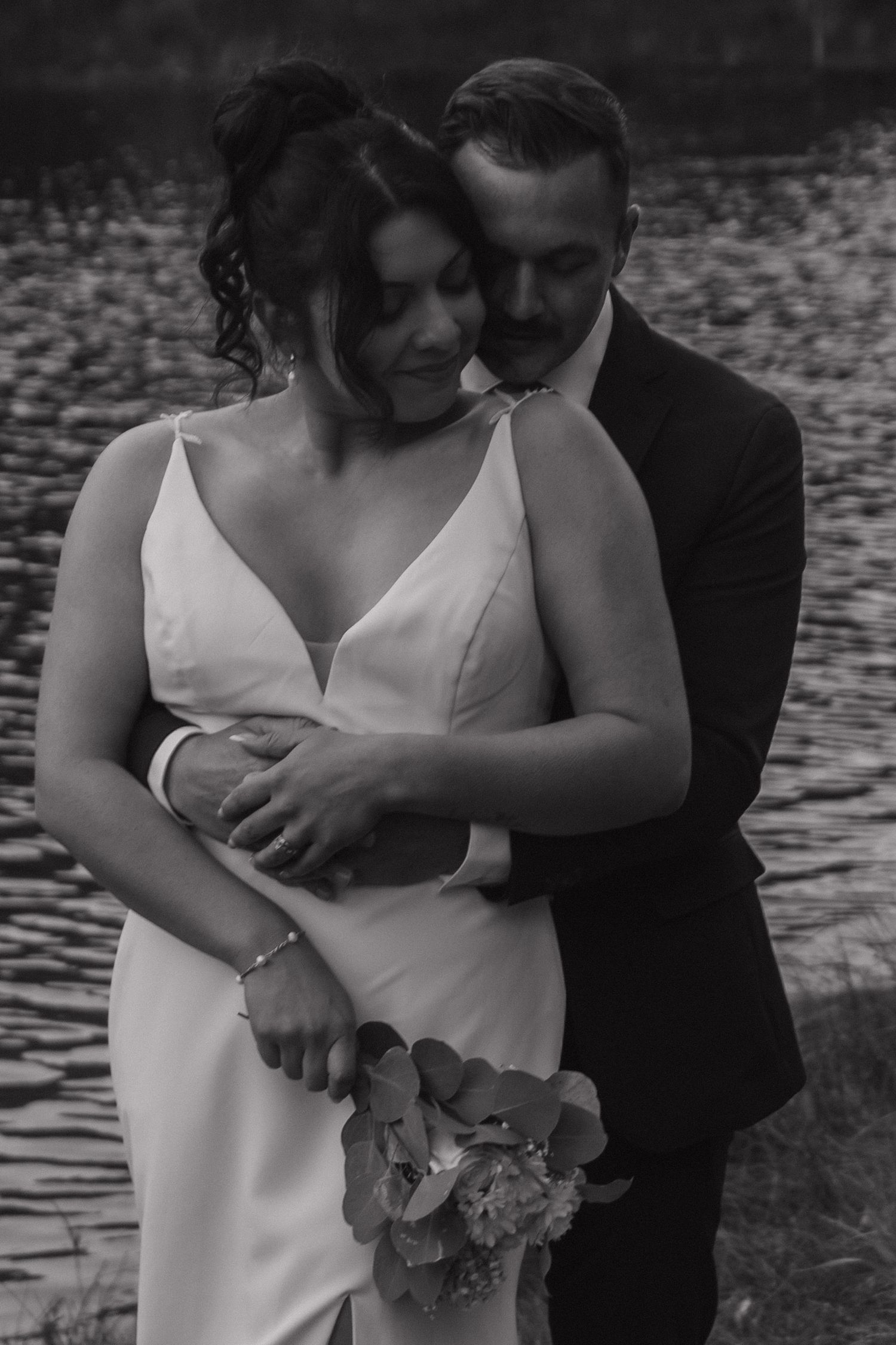 An intimate black and white wedding photo of a couple embracing by a lake.