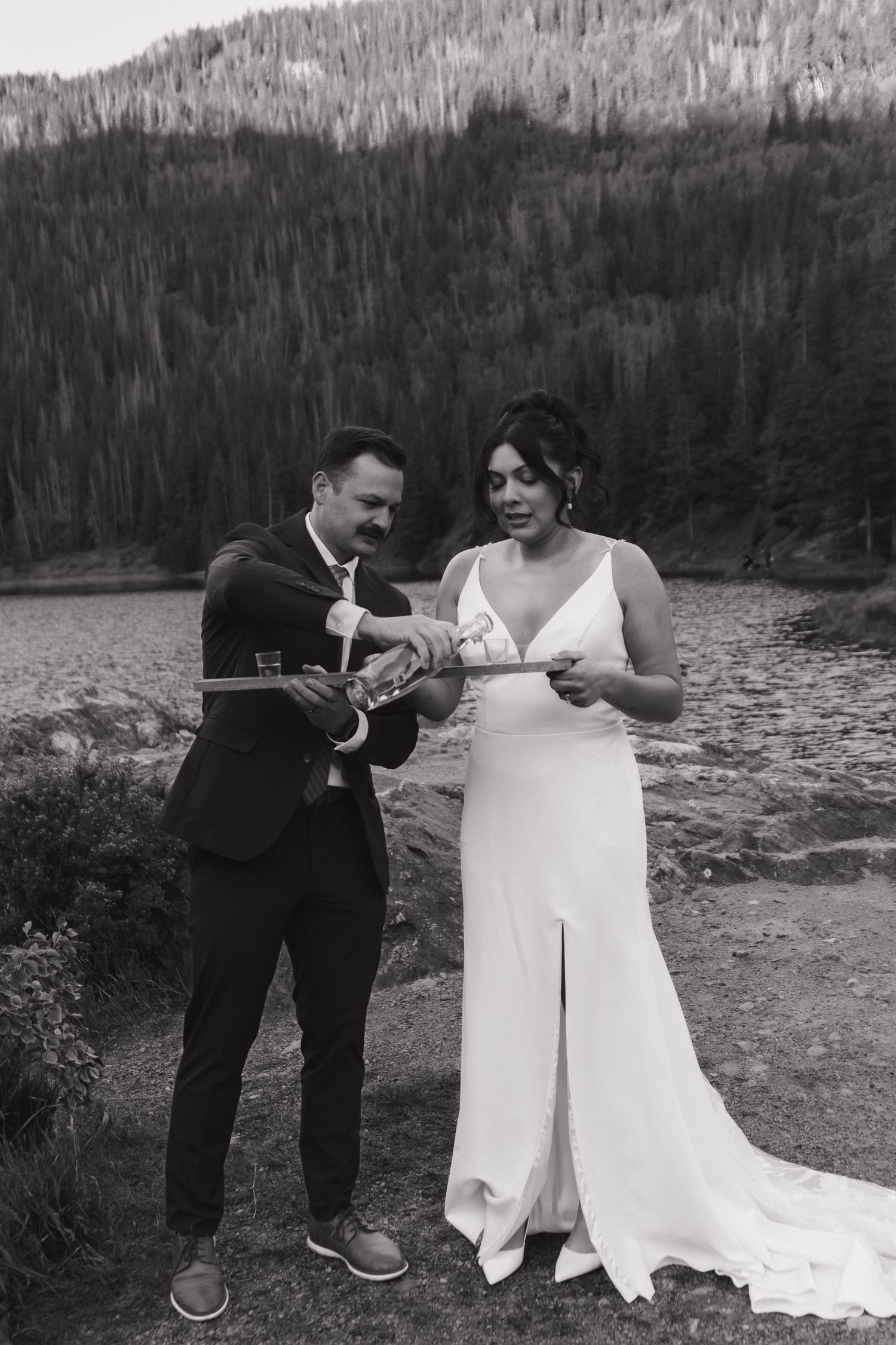 Two people in wedding attire sharing a playful moment near a mountain lake.